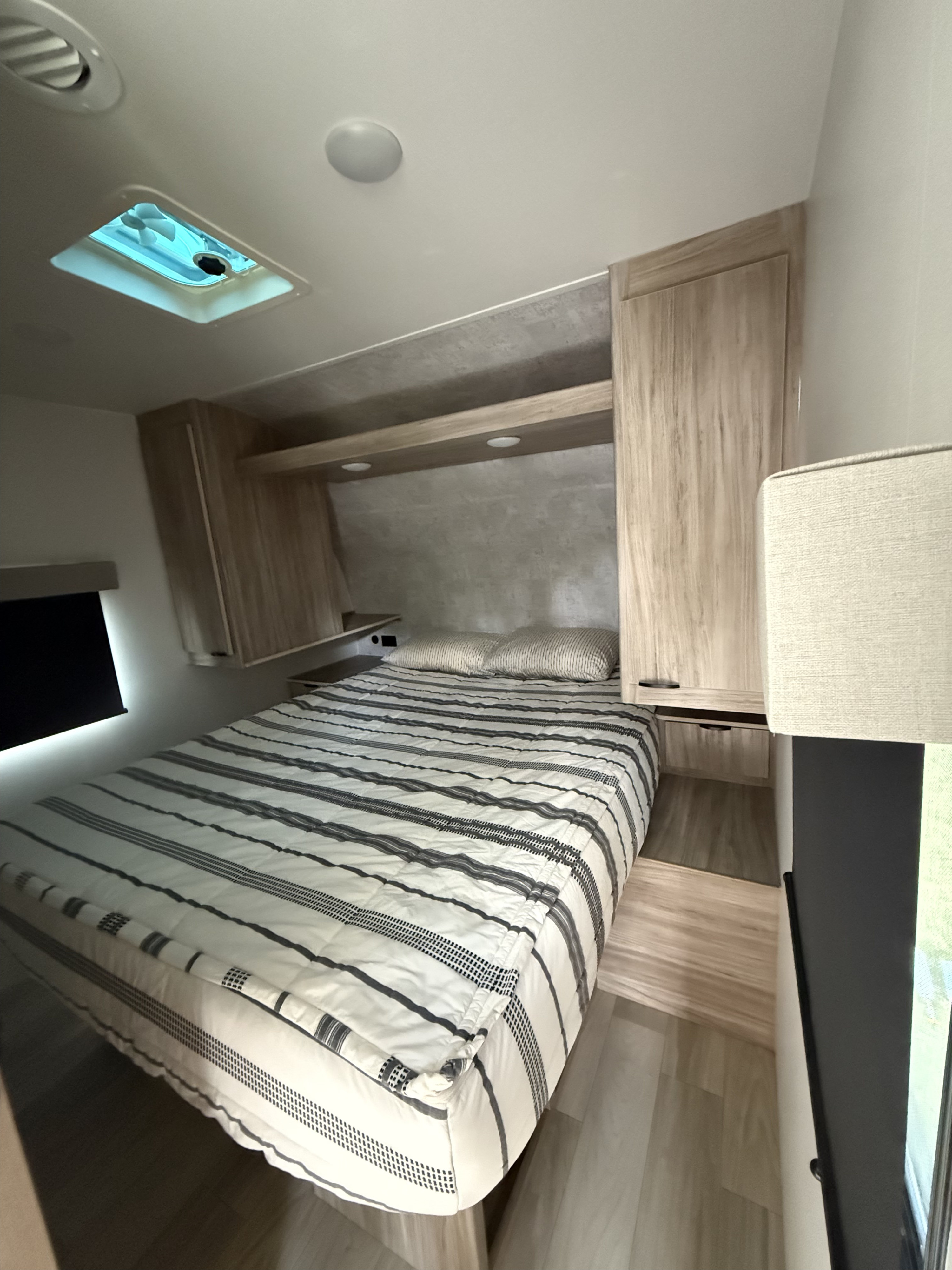 Inside an RV rental: a bright bedroom with striped bedding, wood cabinets, ceiling vent, and wood-look flooring.. Winnebago Access 2024