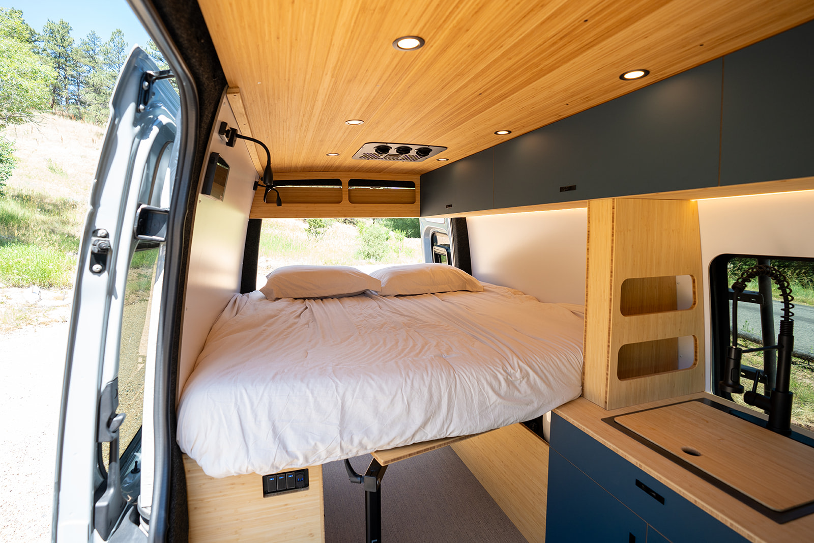 Cozy rv rental van interior: a large bed, sleek bamboo ceiling with lights, and a compact kitchen. Nature visible outside.. Geotrek Albatross Flatiron 2025
