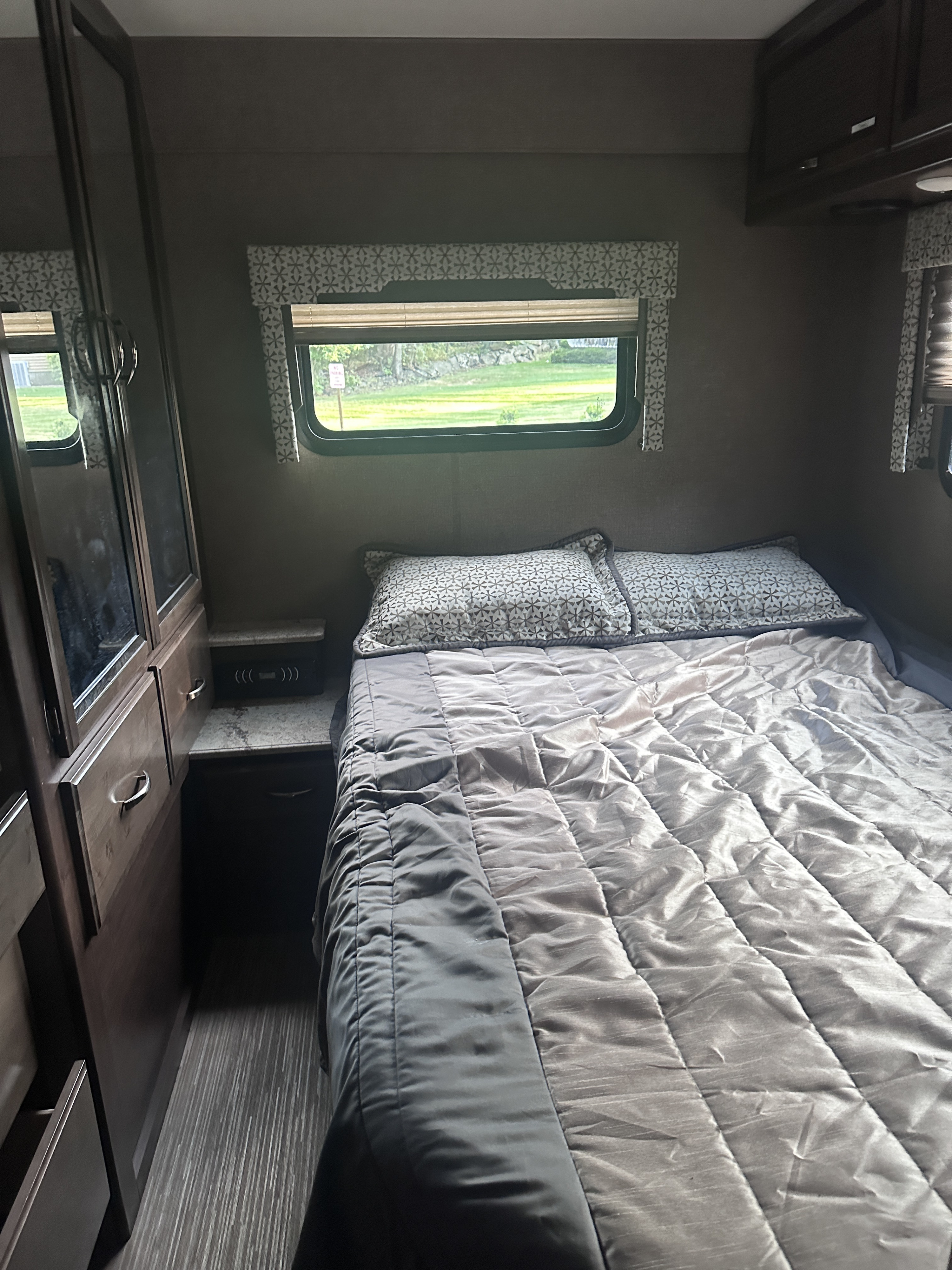 Cozy RV rental bedroom with a bed featuring patterned pillows, window with greenery view, and wood cabinetry.. Thor Axis 2018