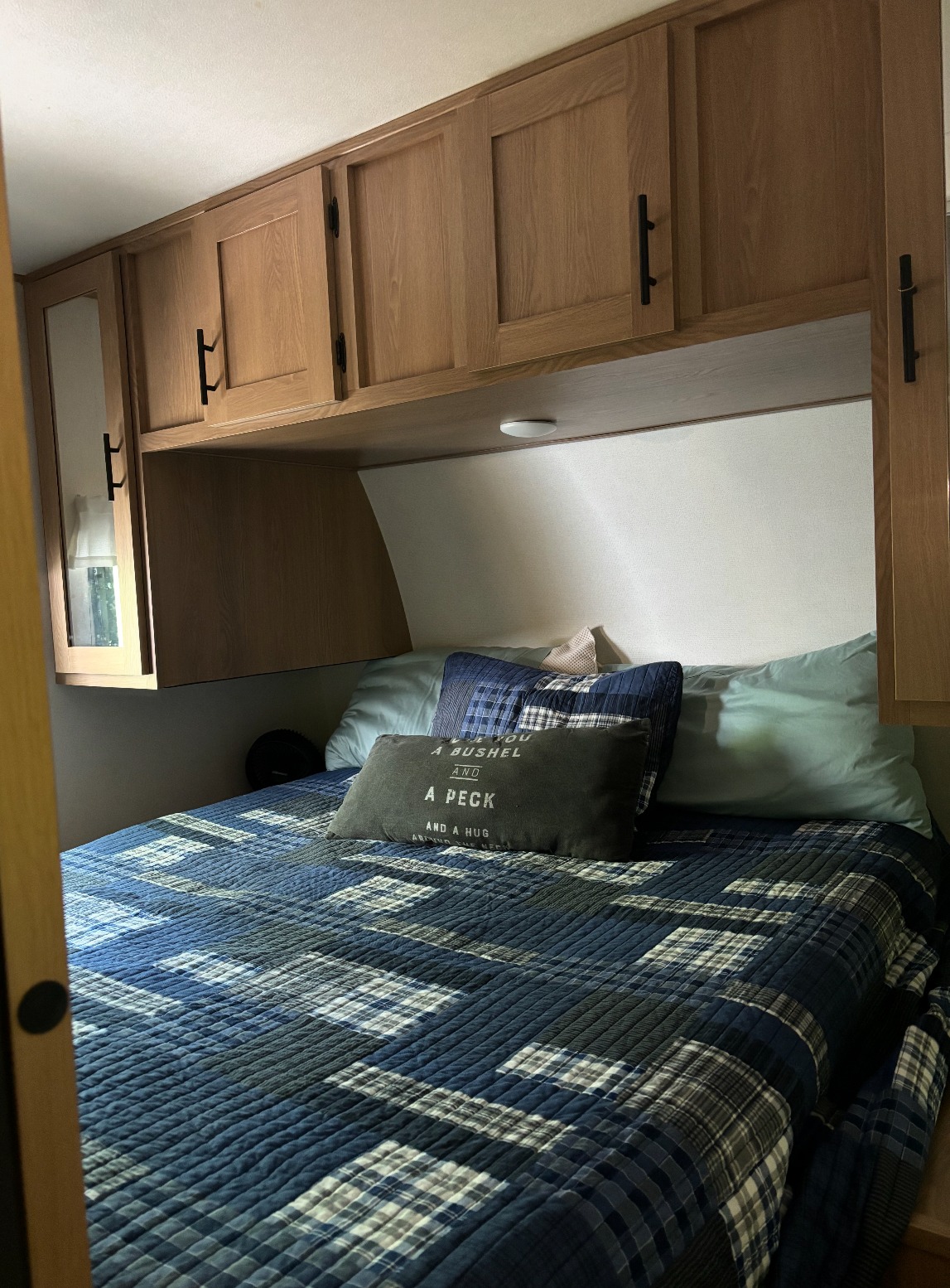 Inside an RV rental: a cozy bedroom with a blue plaid bedspread and overhead wooden cabinets.. Avenger 24BHS 2022