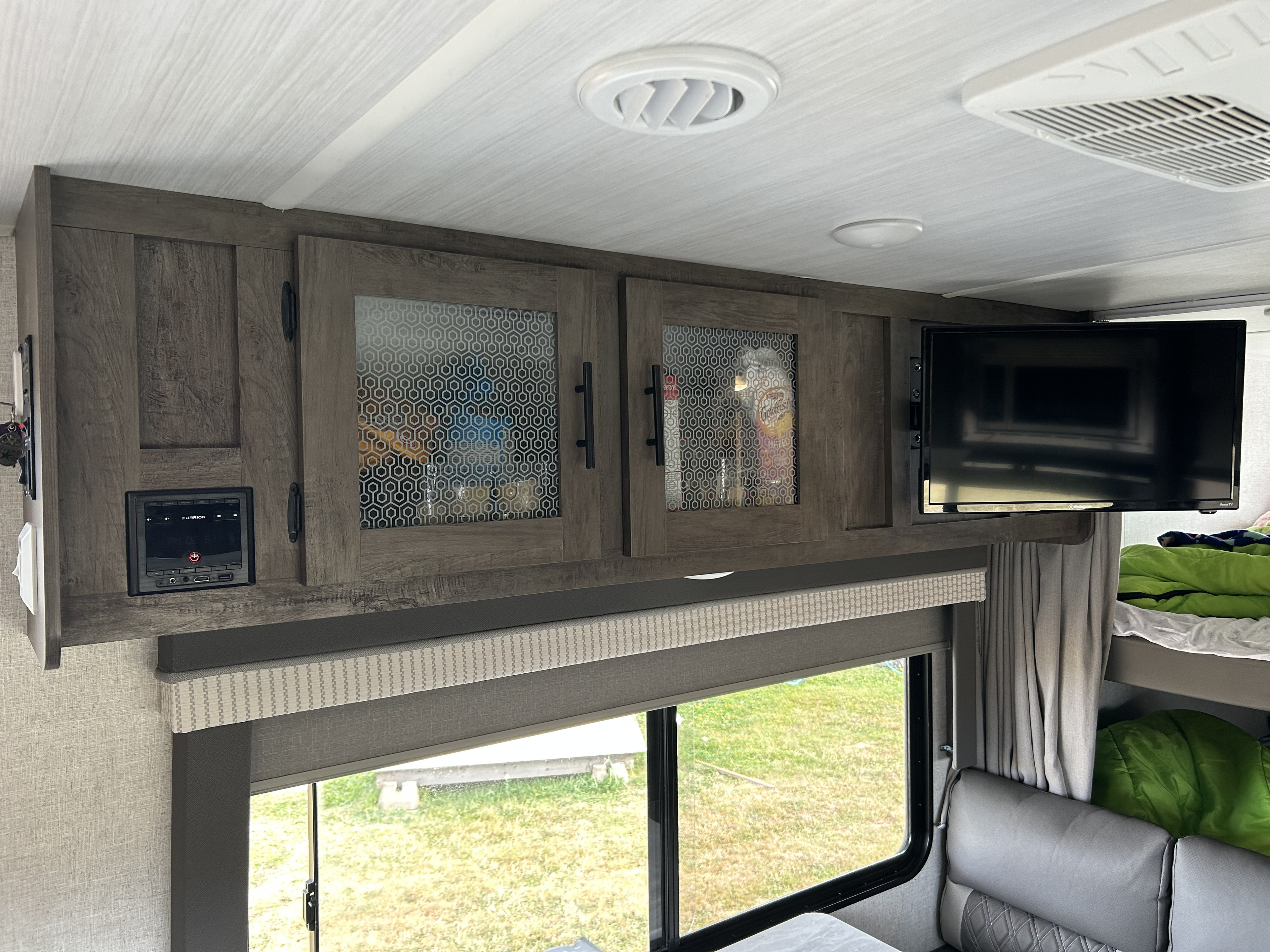 Inside an RV rental: overhead wood cabinets with patterned glass, a TV, a Furrion media player, and a window with blinds.. Forest River Wildwood X-Lite 2021
