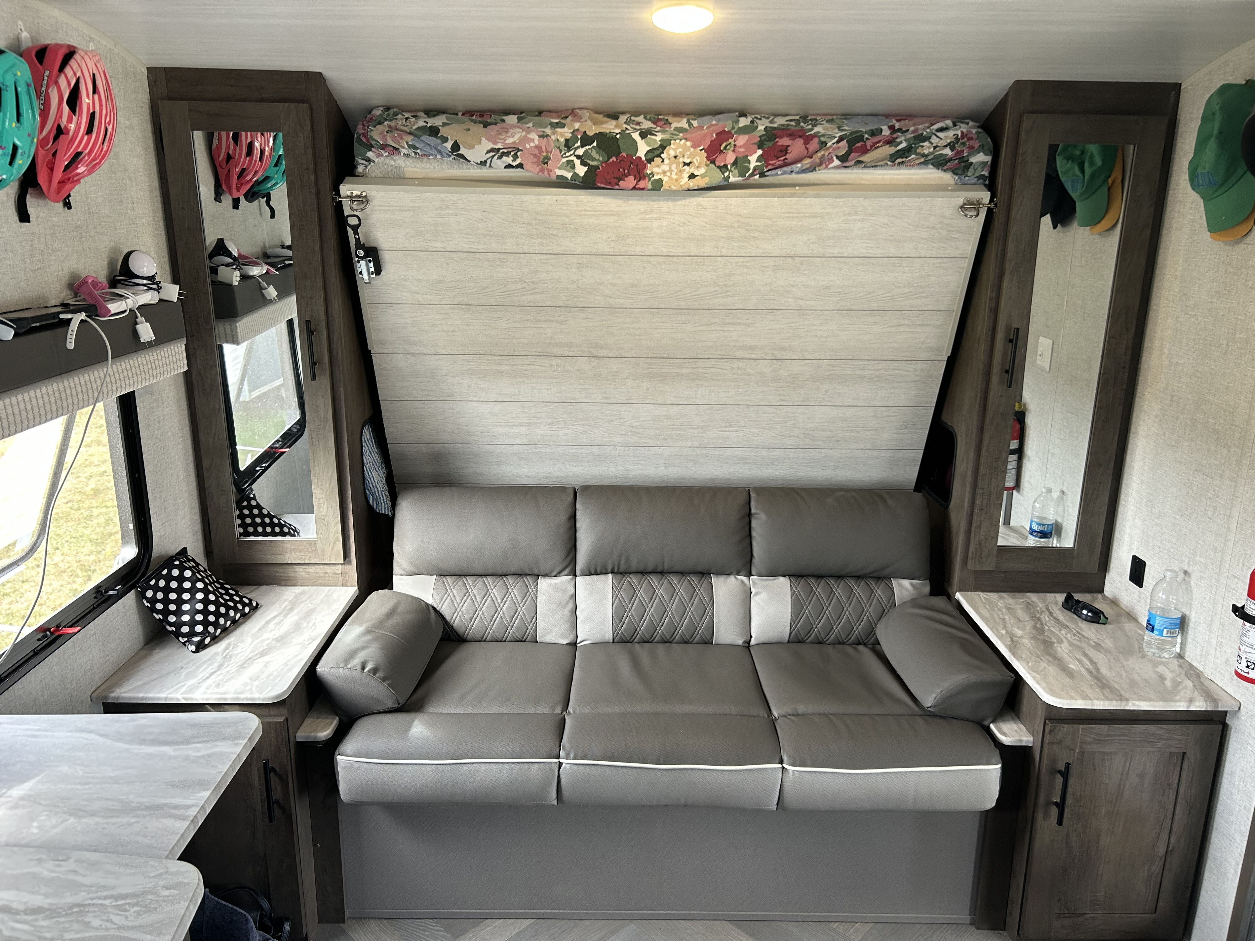Functional RV rental interior with a gray sofa, pull-down bed, and side storage. Helmets and charging station visible.. Forest River Wildwood X-Lite 2021