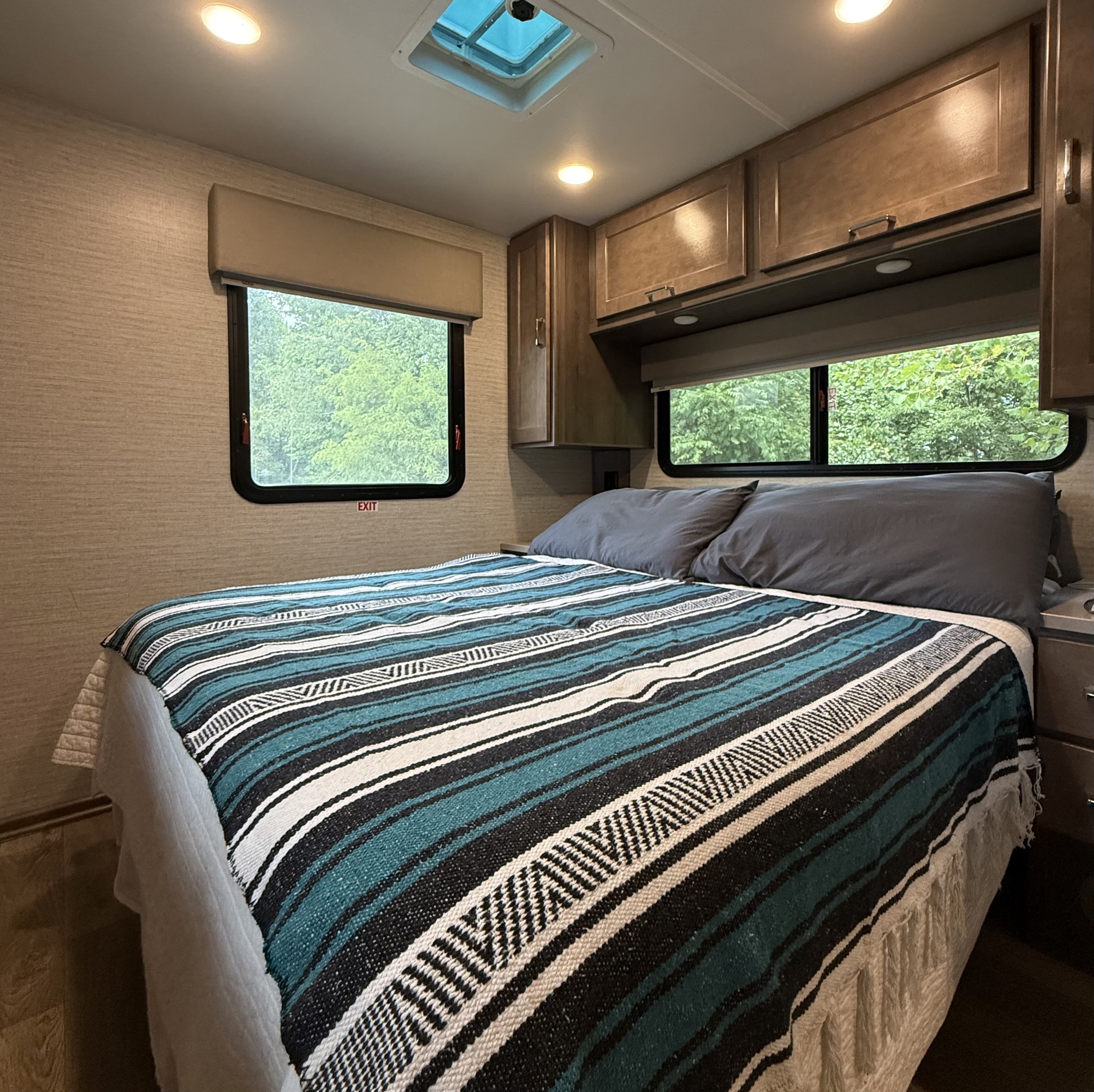 Comfy RV rental bedroom with striped blanket, overhead cabinets, and windows showing green trees outside, perfect for travel adventures.. Winnebago Minnie Winnie 2021