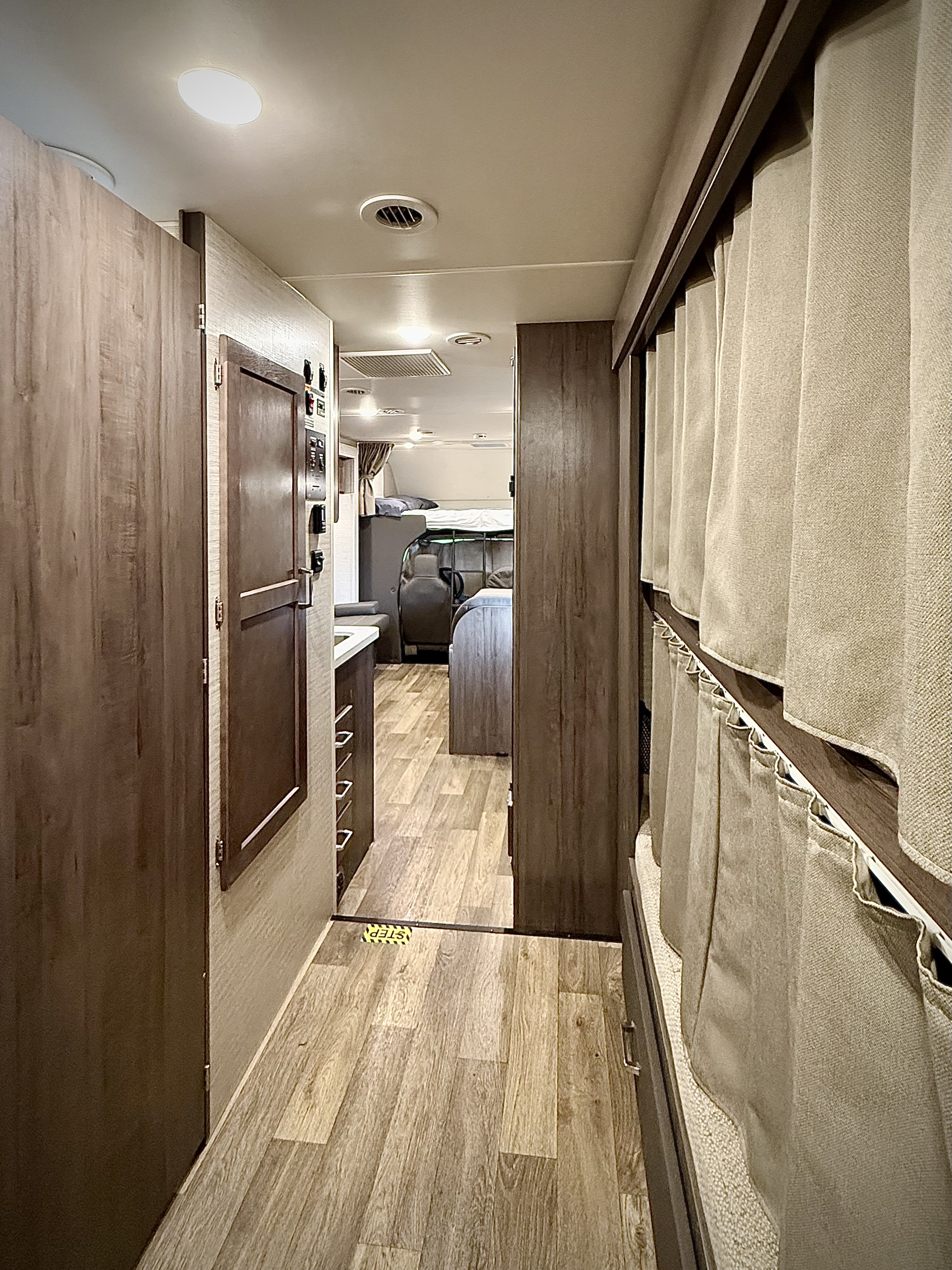 View down the central aisle of an RV rental, revealing a kitchen area, bunk beds with privacy curtains, and various controls.. Winnebago Minnie Winnie 2021