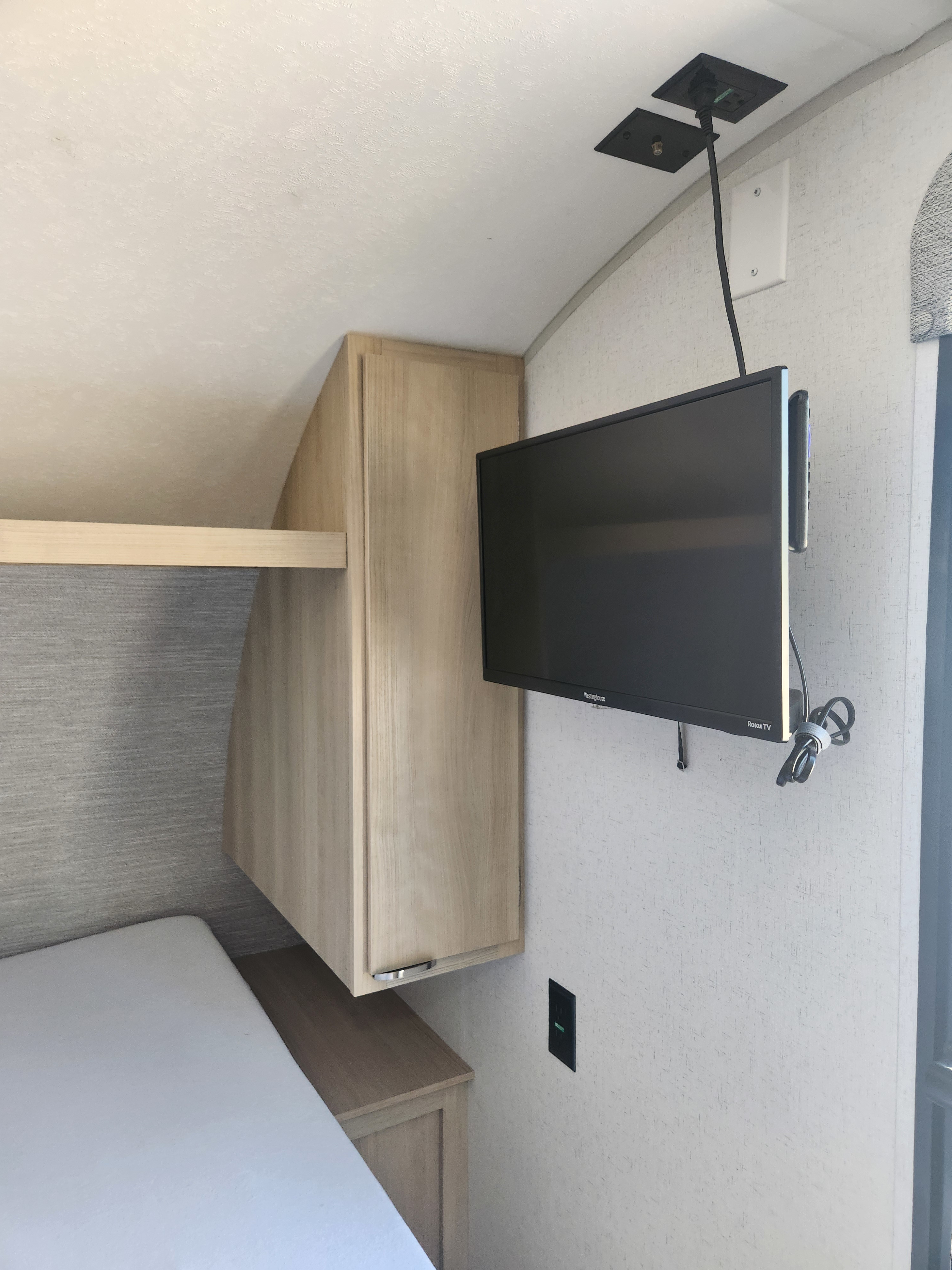 Compact RV rental bedroom showing a bed, wall-mounted Westinghouse Roku TV, and light wood cabinetry for efficient space.. Forest River Wildwood FSX 174BHLE 2024