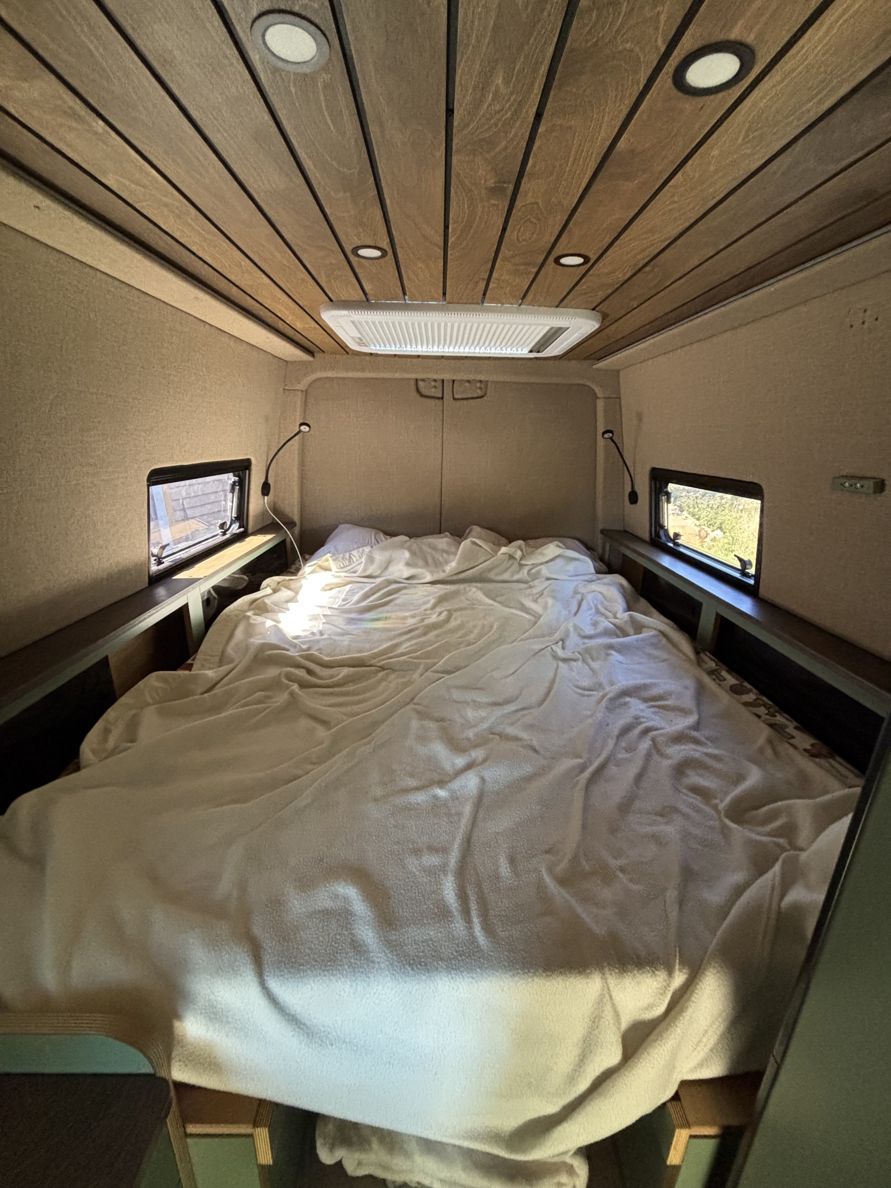 Cozy RV rental van interior showcasing a large, inviting bed, side windows, and wood-paneled ceiling for comfortable sleep.. Mercedes-Benz Sprinter 2025