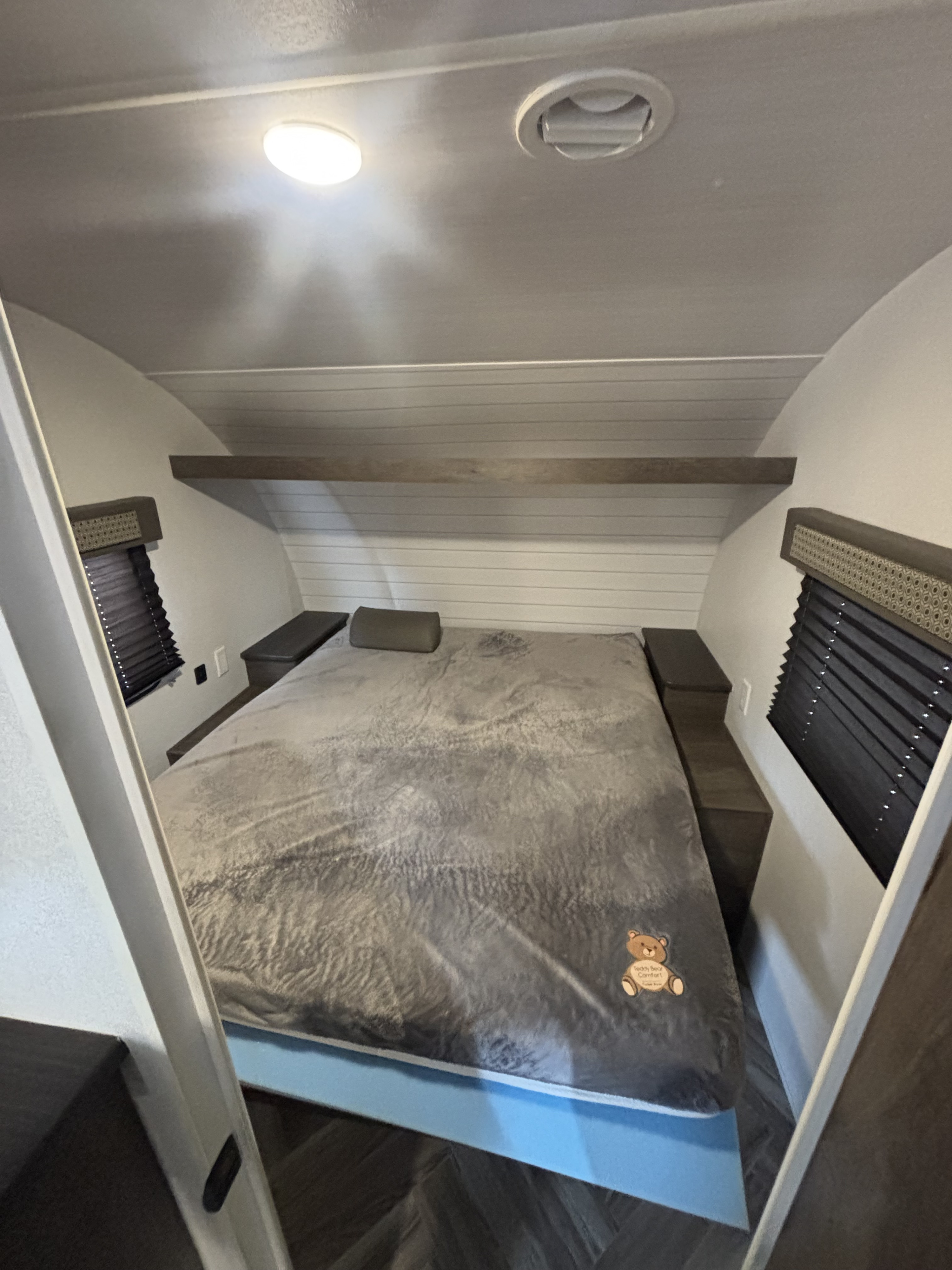 Explore this compact RV rental bedroom: a cozy bed with grey comforter, built-in nightstands, and window blinds.. Forest River Wildwood X-Lite 2023