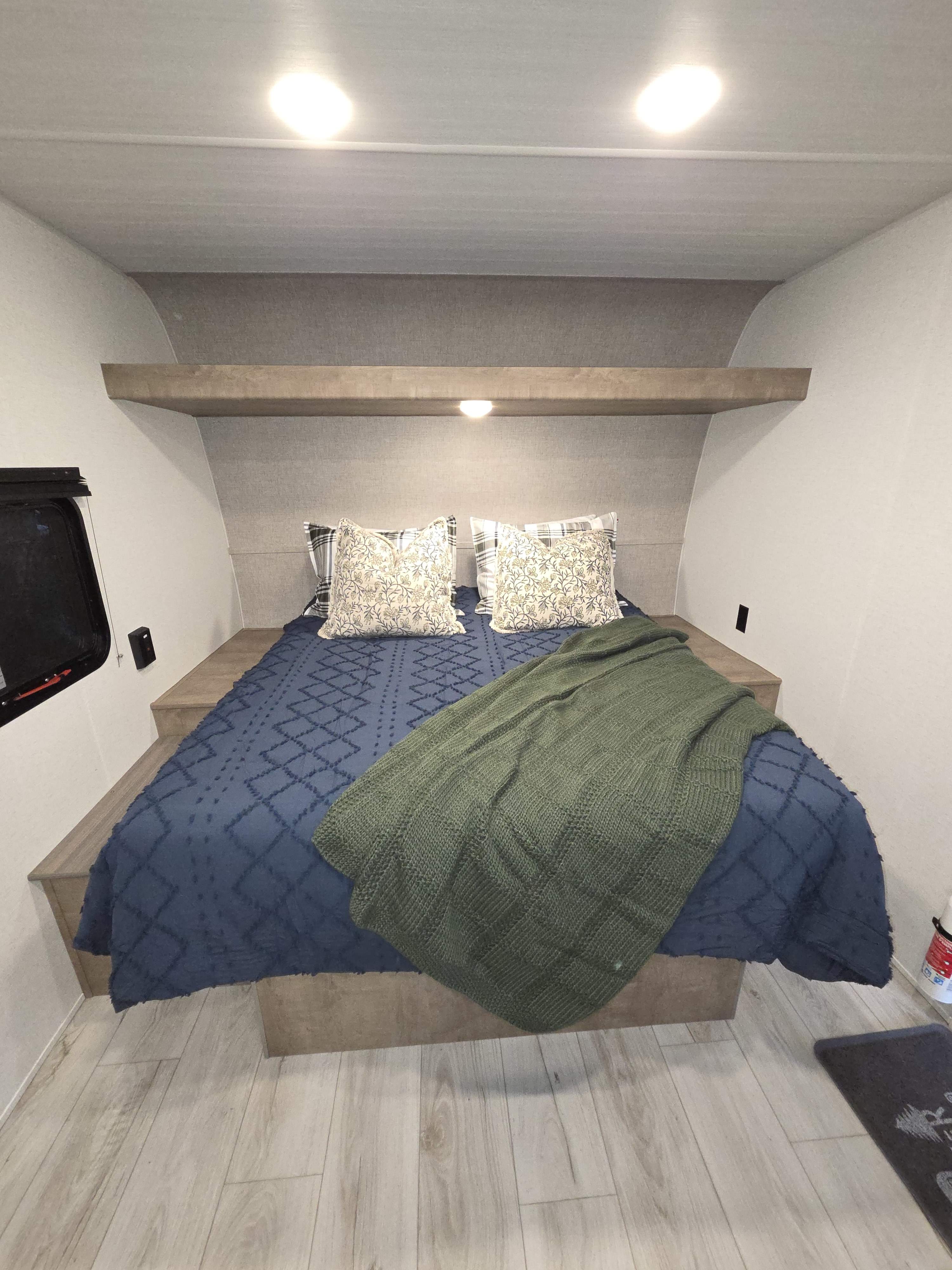 Cozy RV rental bedroom with a blue bedspread, green blanket, and decorative pillows. Features overhead shelf & window.. Keystone Springdale West BHWE 2025