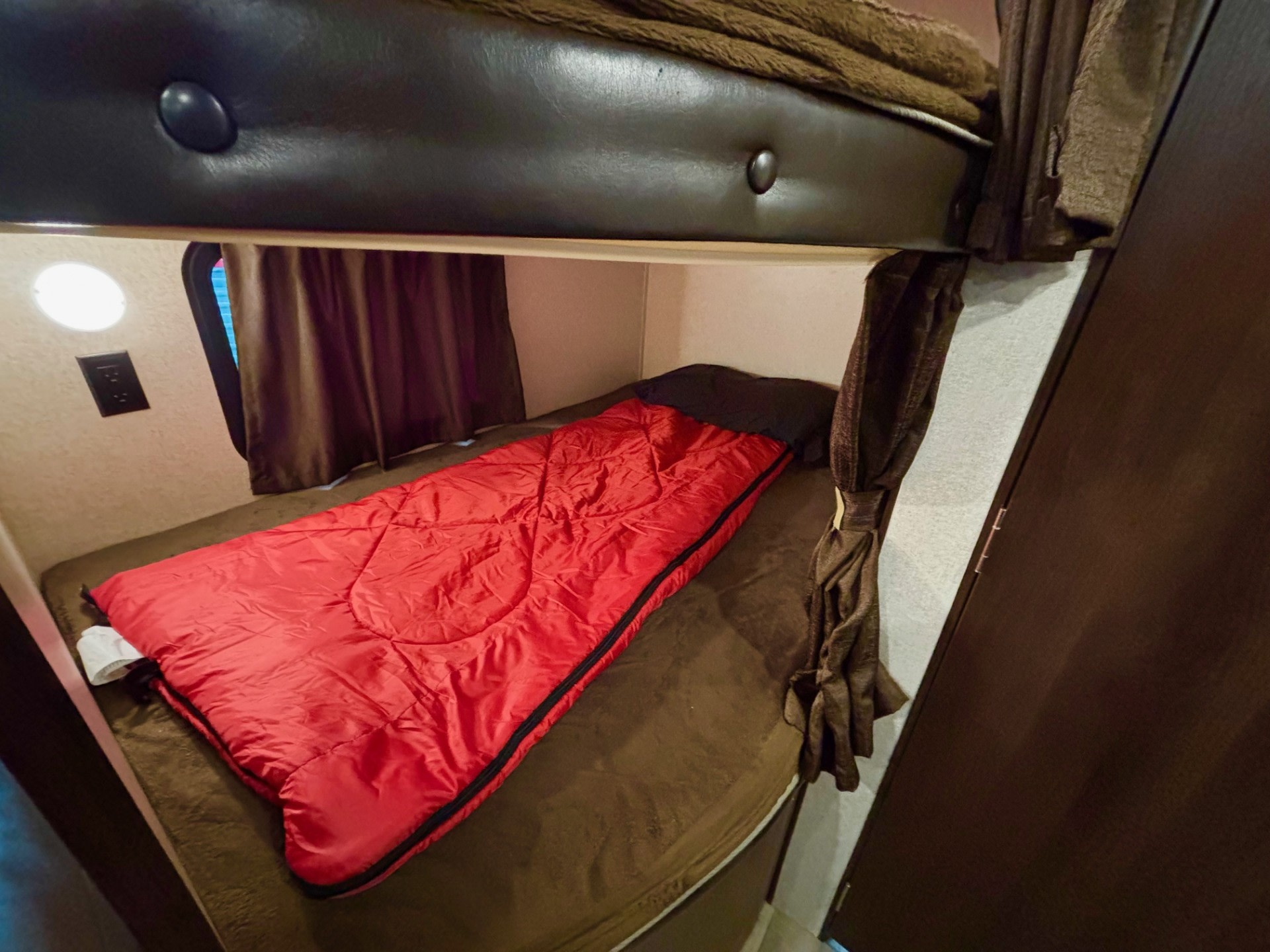 RV rental interior: A lower bunk bed with a bright red sleeping bag and black pillow, framed by dark brown curtains.. Jayco Jay Flight 2019
