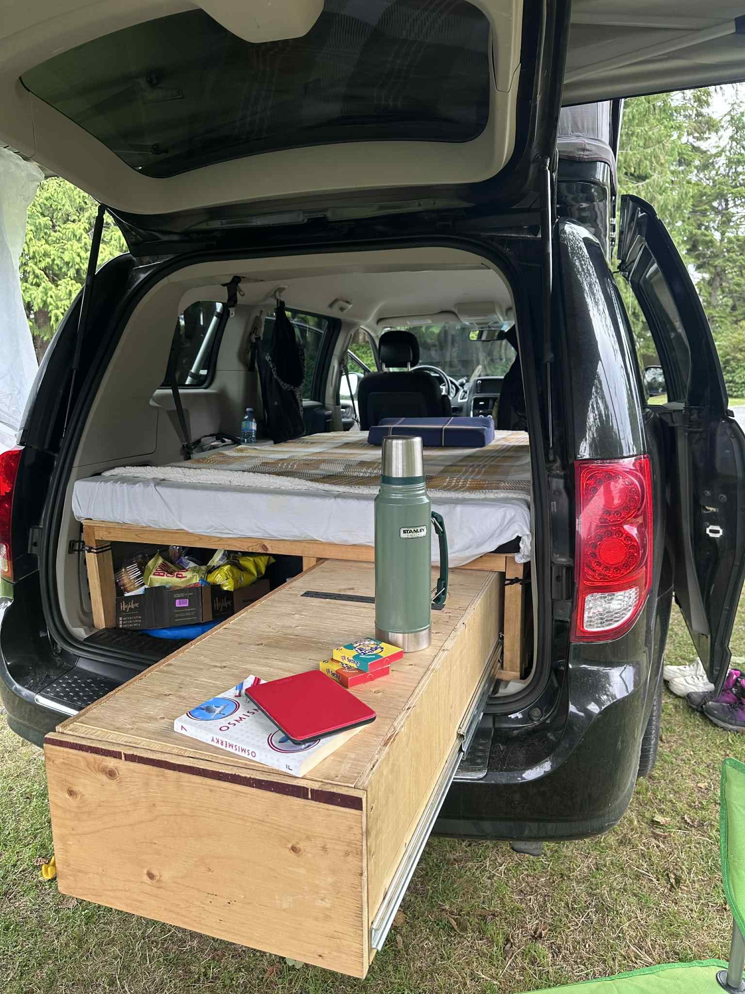 Open tailgate of a black minivan rv rental, revealing a camping bed, storage, and extended wooden table.. Dodge Grand Caravan 2014