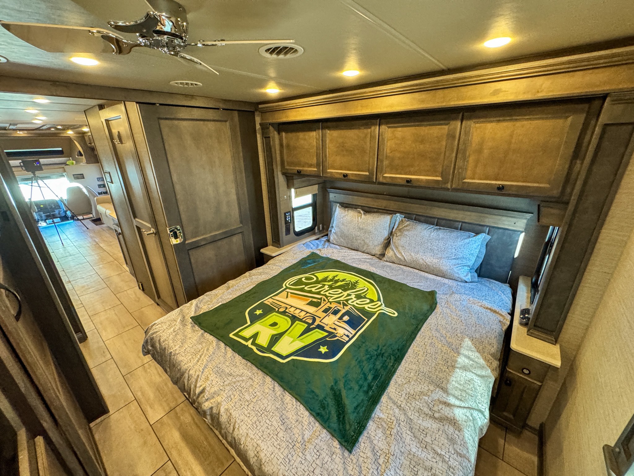 Inside a luxurious RV rental bedroom with a large bed, ceiling fan, and overhead storage. Tiled flooring leads to the front.. Tiffin Allegro Bay Super-C 2022