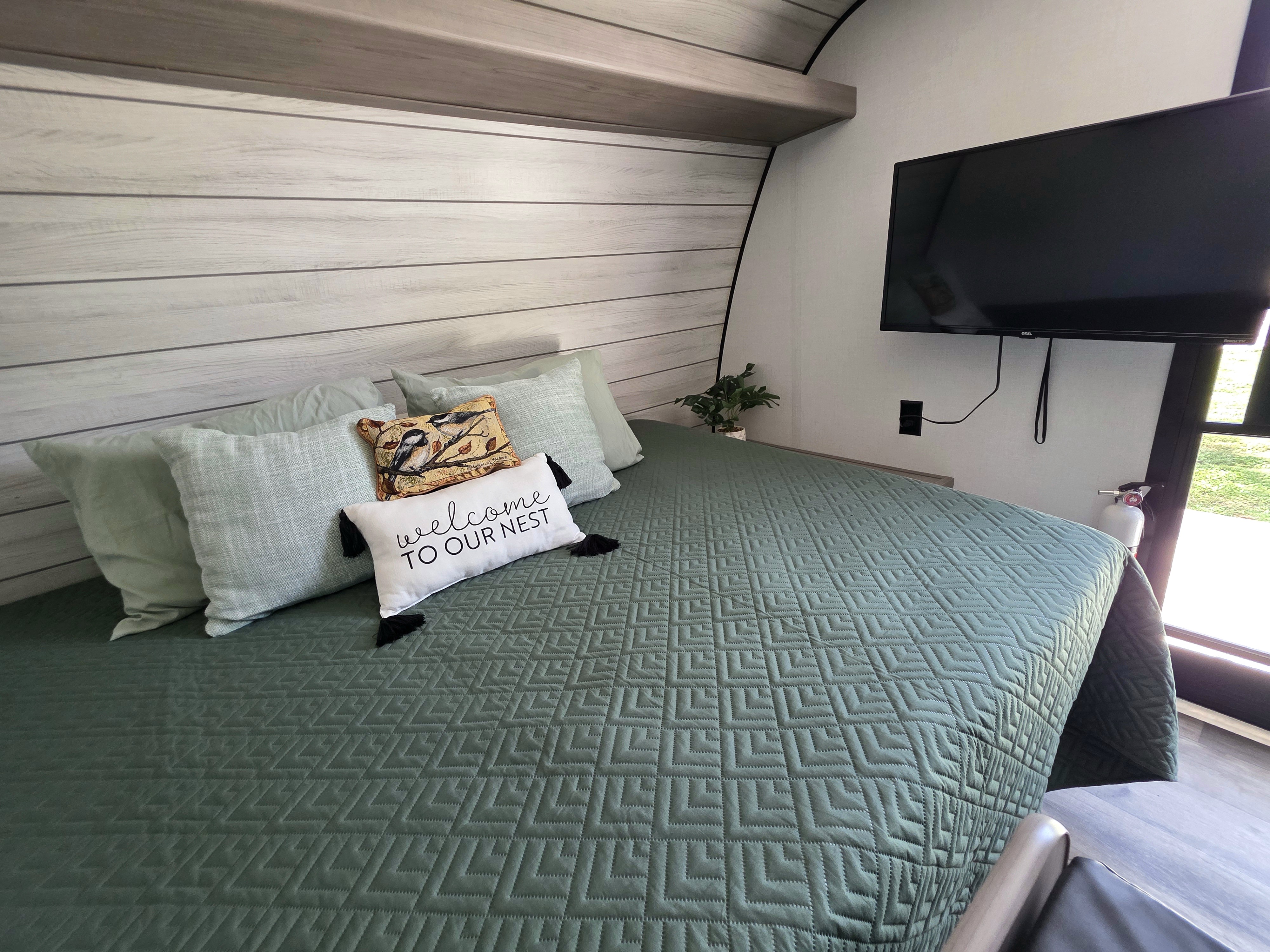 Relax in this cozy RV rental bedroom. Features a green quilted bed, "welcome" pillow, light wood walls, and a TV.. Dutchman Coleman 2024