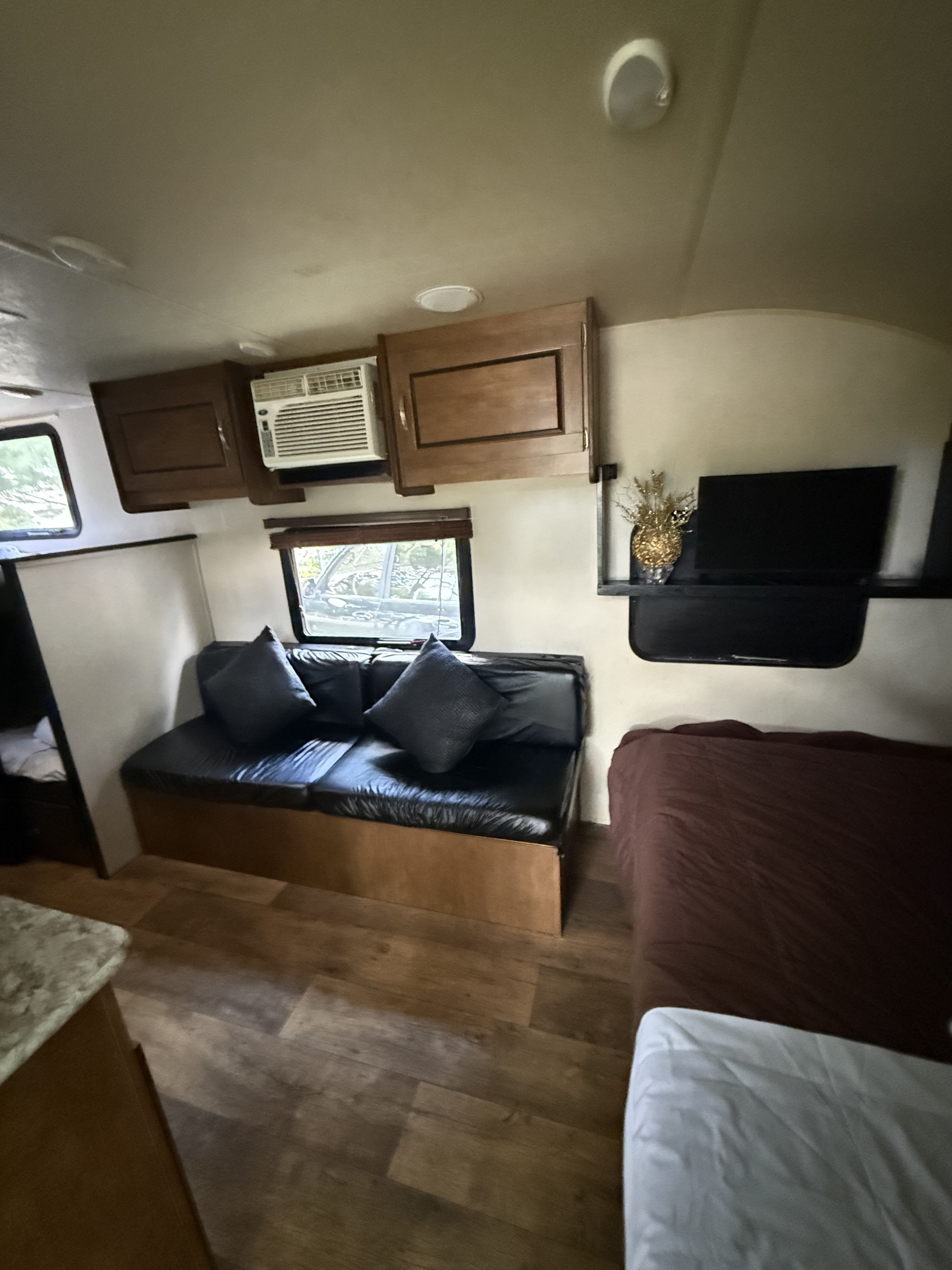 Inside an RV rental: view of living area with black couch, windows, wall AC, TV, and cozy bed. Wood-look floors.. Forest River Salem Cruise Lite 2019