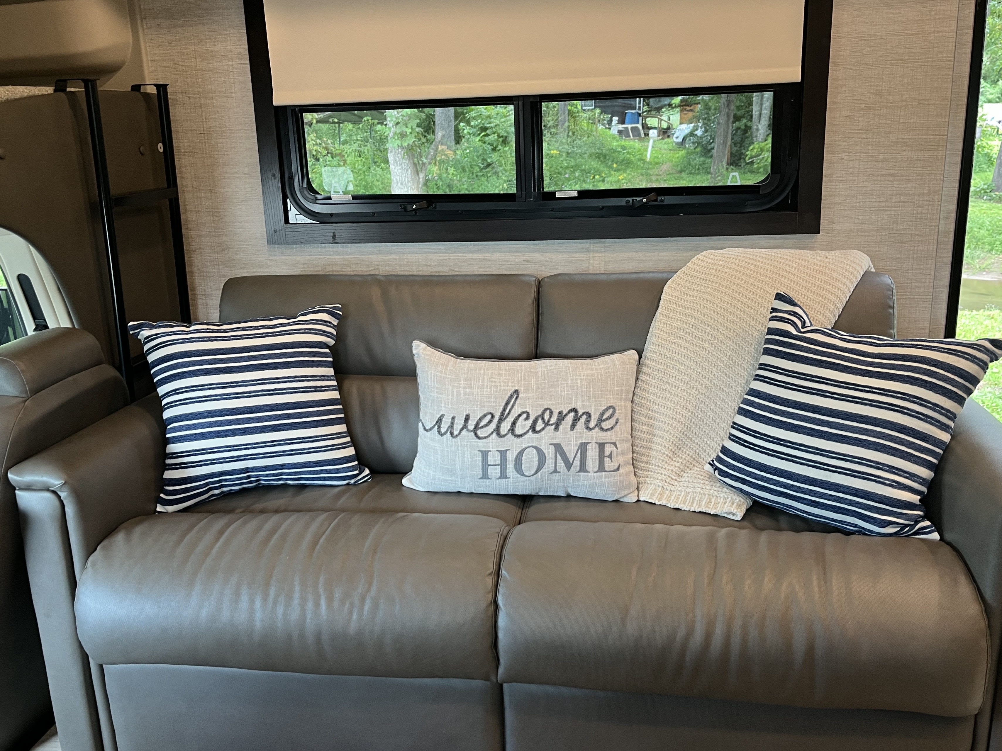 Cozy RV rental sofa with "welcome HOME" and striped pillows, plus a cream throw, overlooking a nature view.. Entegra Coach Esteem 2021