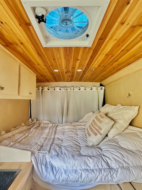 Cozy RV rental interior showing a comfortable bed, wood-paneled ceiling with a fan, and white curtains.. Dodge Ram Promaster 2014