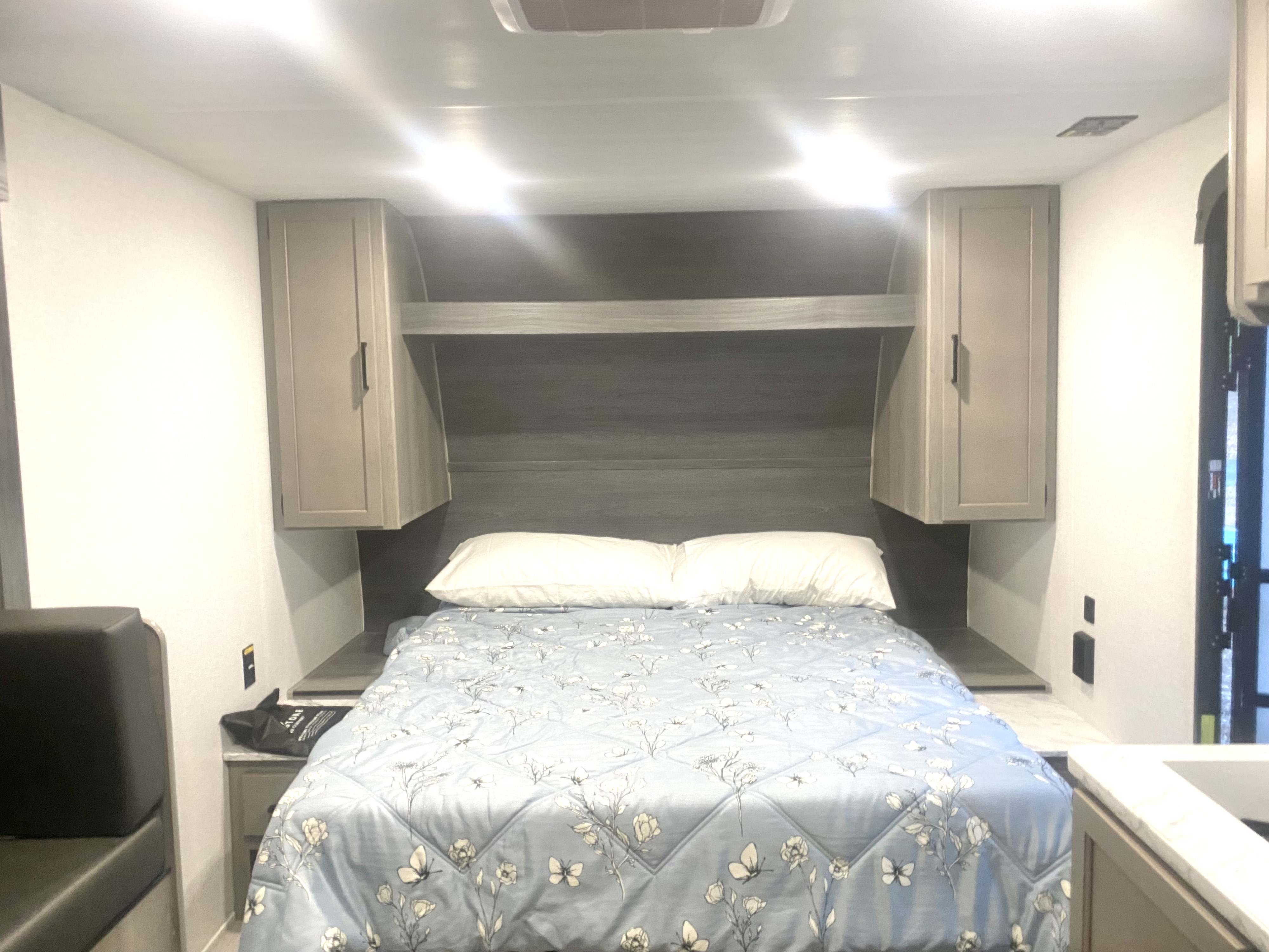 Cozy RV rental bedroom featuring a bed with blue floral comforter, white pillows, overhead cabinets, and bright lights.. Keystone RV Hideout Sport 2024