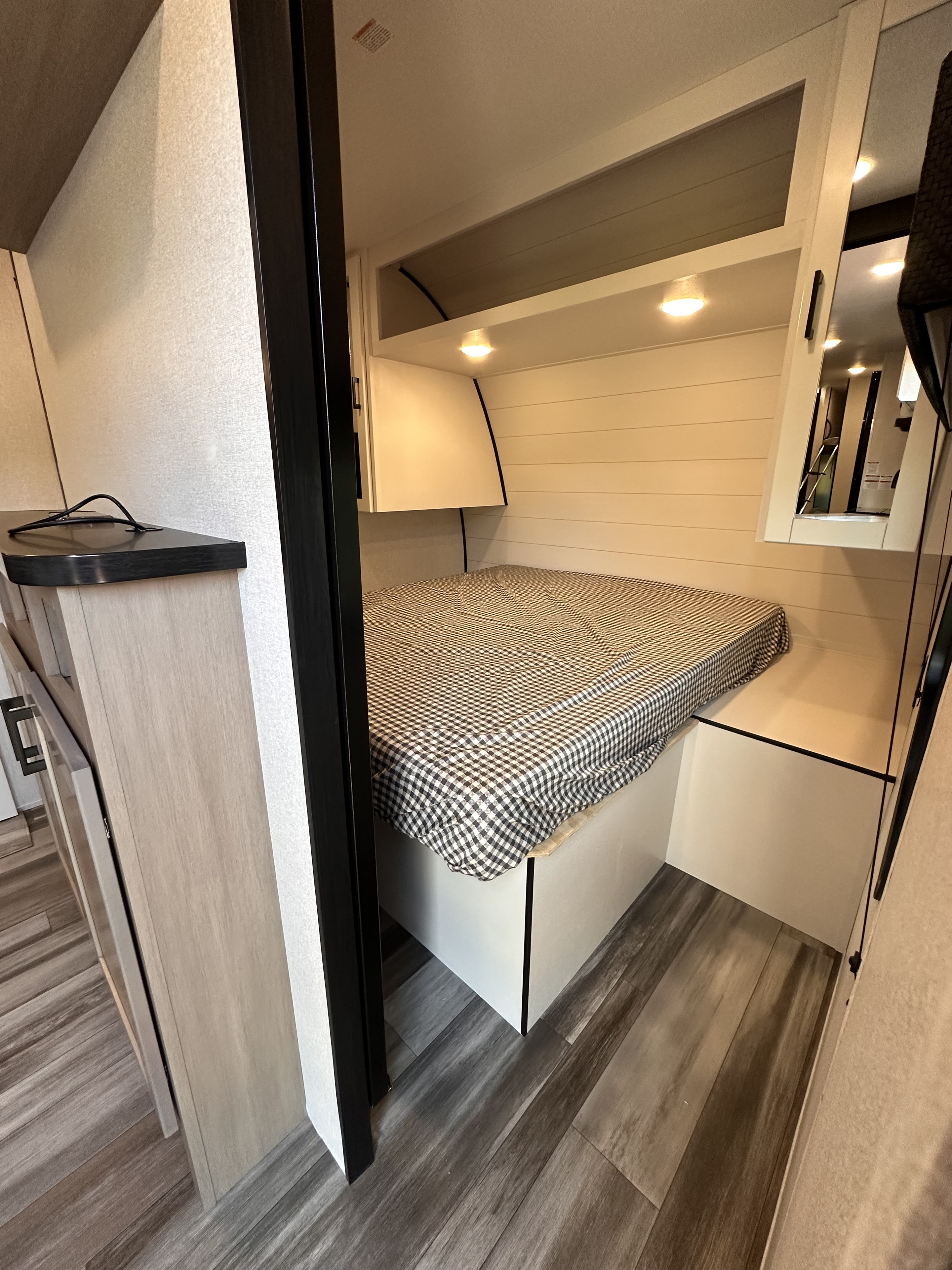 Compact RV rental sleeping area with checkered bed, shiplap walls, overhead lights, and built-in storage.. Jayco Jay Flight 2026