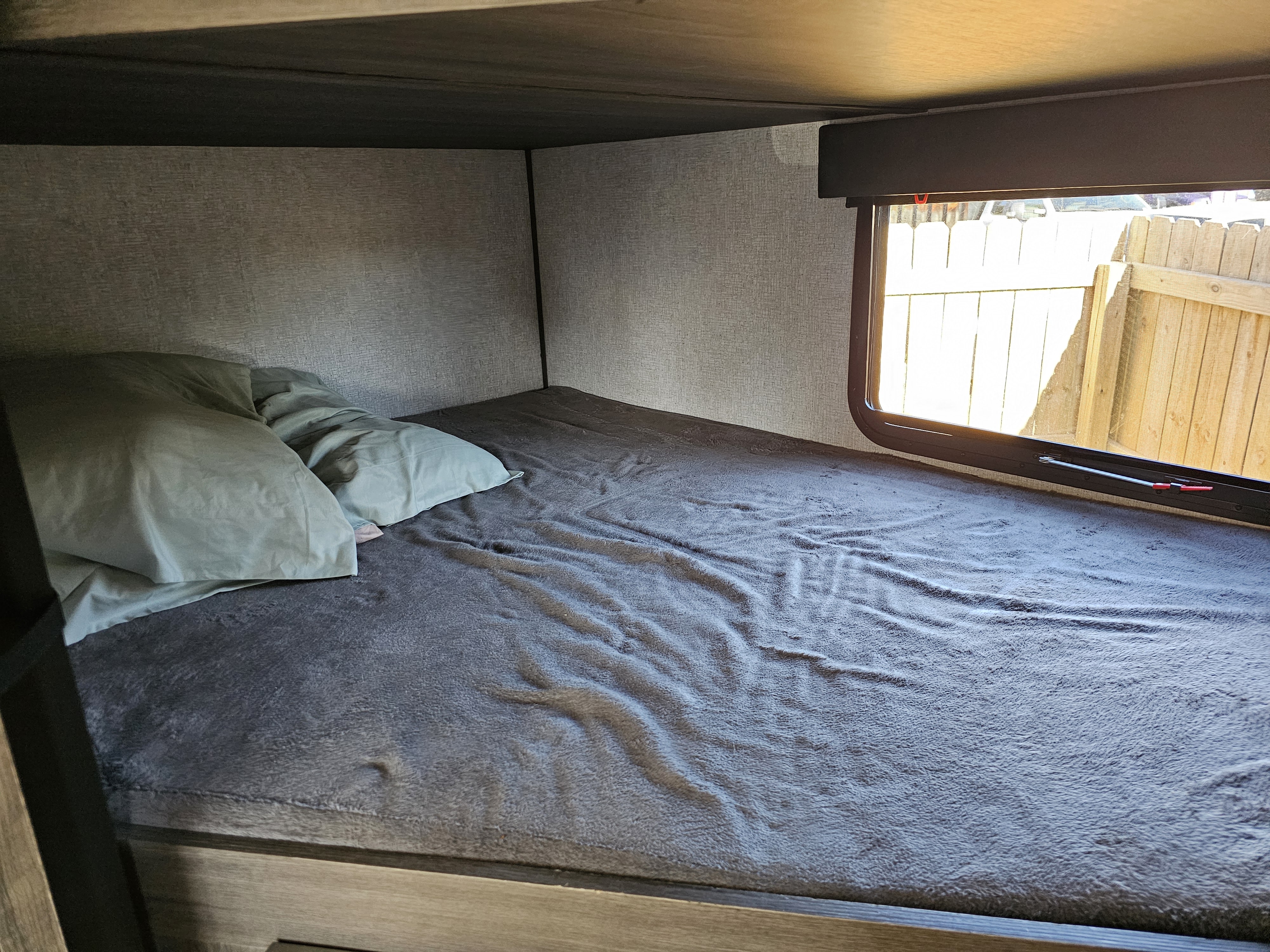 An RV rental's cozy bunk bed, featuring soft gray bedding, green pillows, and a window view of a wooden fence.. Dutchmen Kodiak Ultra Lite 2021