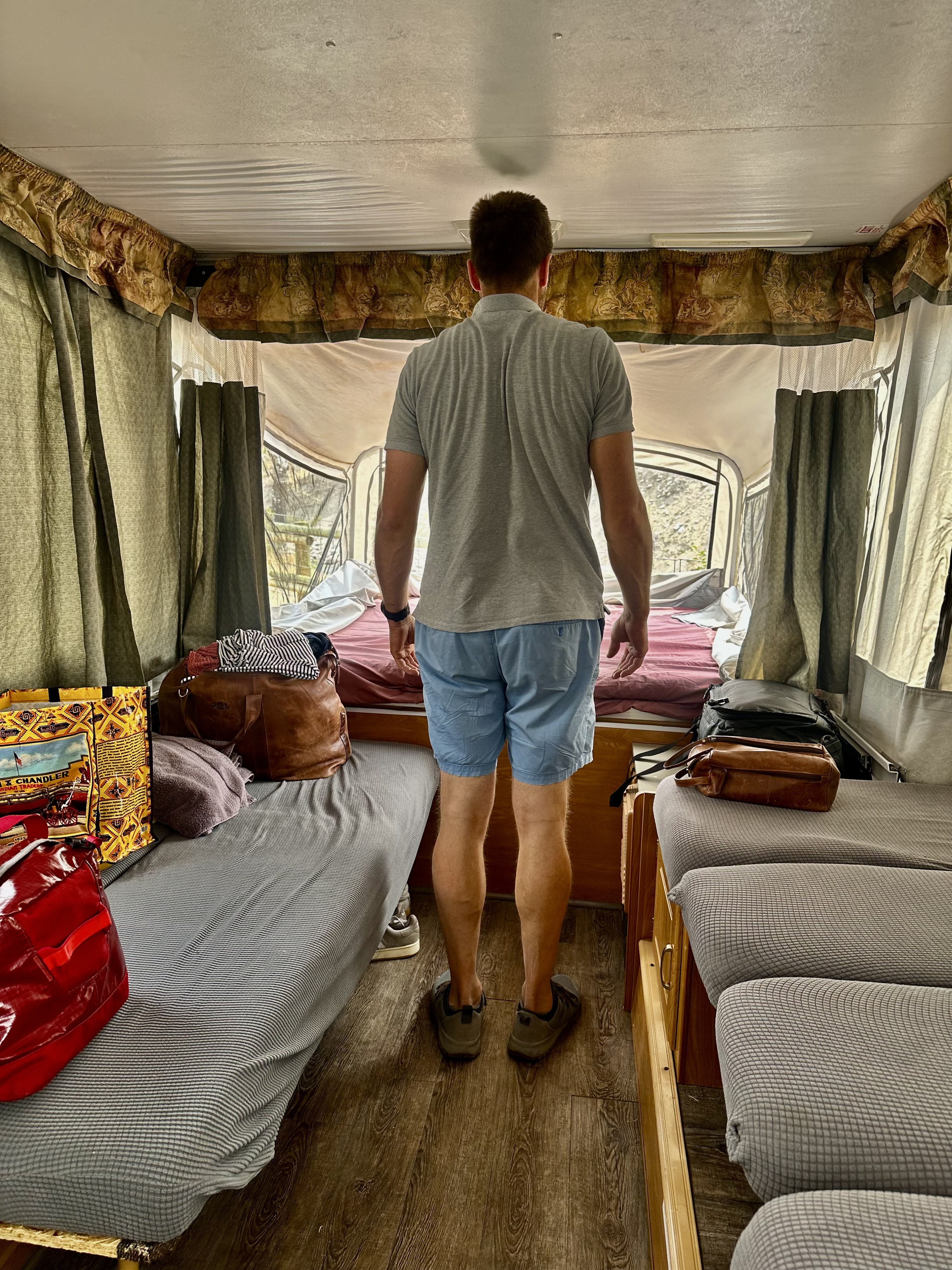 Inside an RV rental, a person stands looking out the back of the pop-up camper at the sleeping area and packed luggage.. Coleman Americana Utah 2004