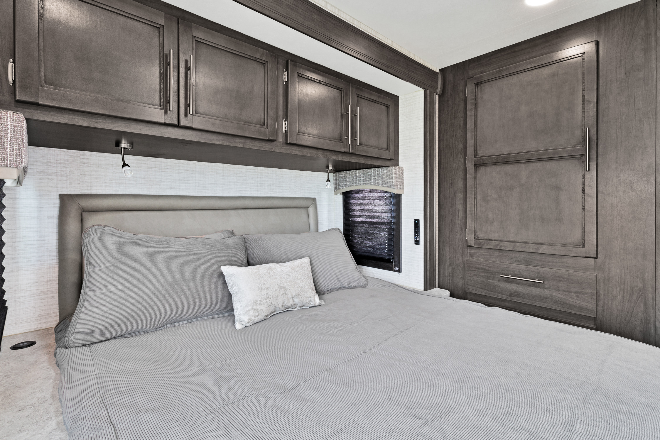 Experience comfort in this modern RV rental bedroom with gray bedding, built-in dark wood cabinets, and integrated lighting.. Jayco Redhawk SE 2024