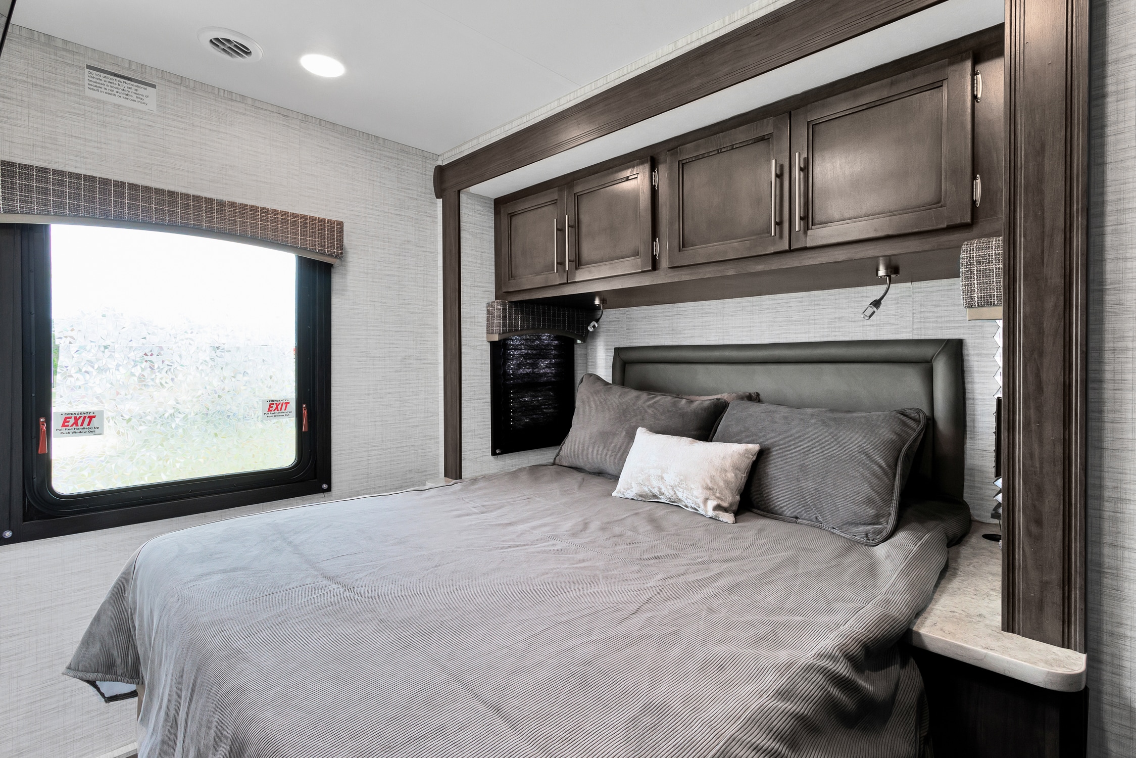 Spacious RV rental bedroom with a grey bed, dark wood overhead cabinets, textured walls, and emergency exit window.. Jayco Redhawk SE 2024