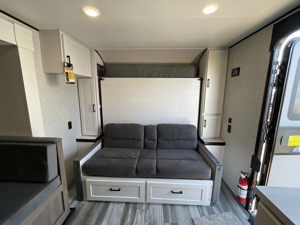 An RV rental's bright interior features a grey sofa that converts to a murphy bed, white cabinets, and an open entry door.. Jayco Jay Flight SLX Baja Edition 2025