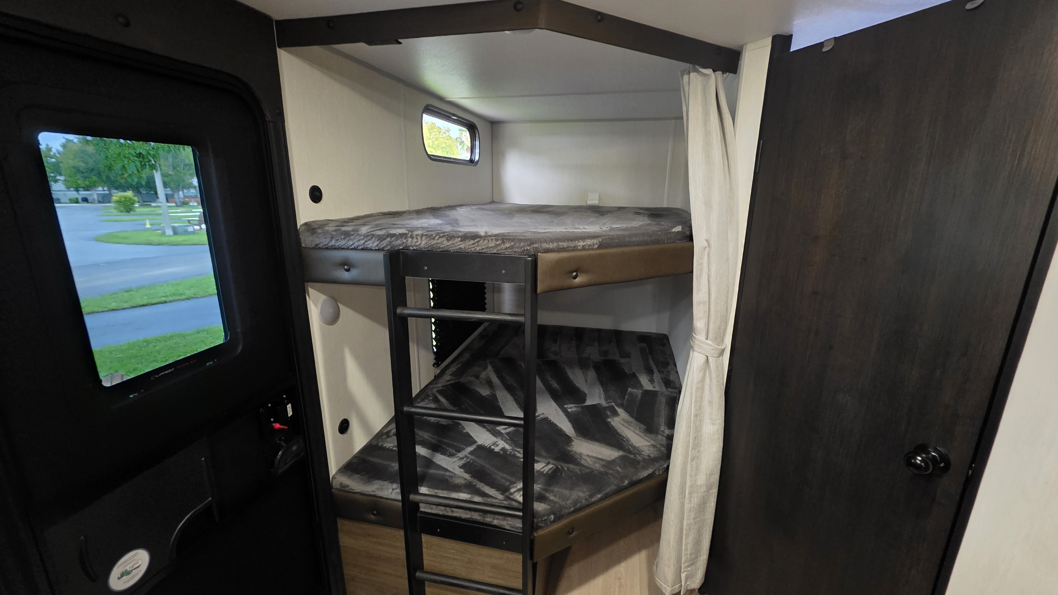 Inside this RV rental, dual bunk beds with a ladder provide compact sleeping. A small window offers a view of a park.. Forest River Cherokee Grey Wolf 2025