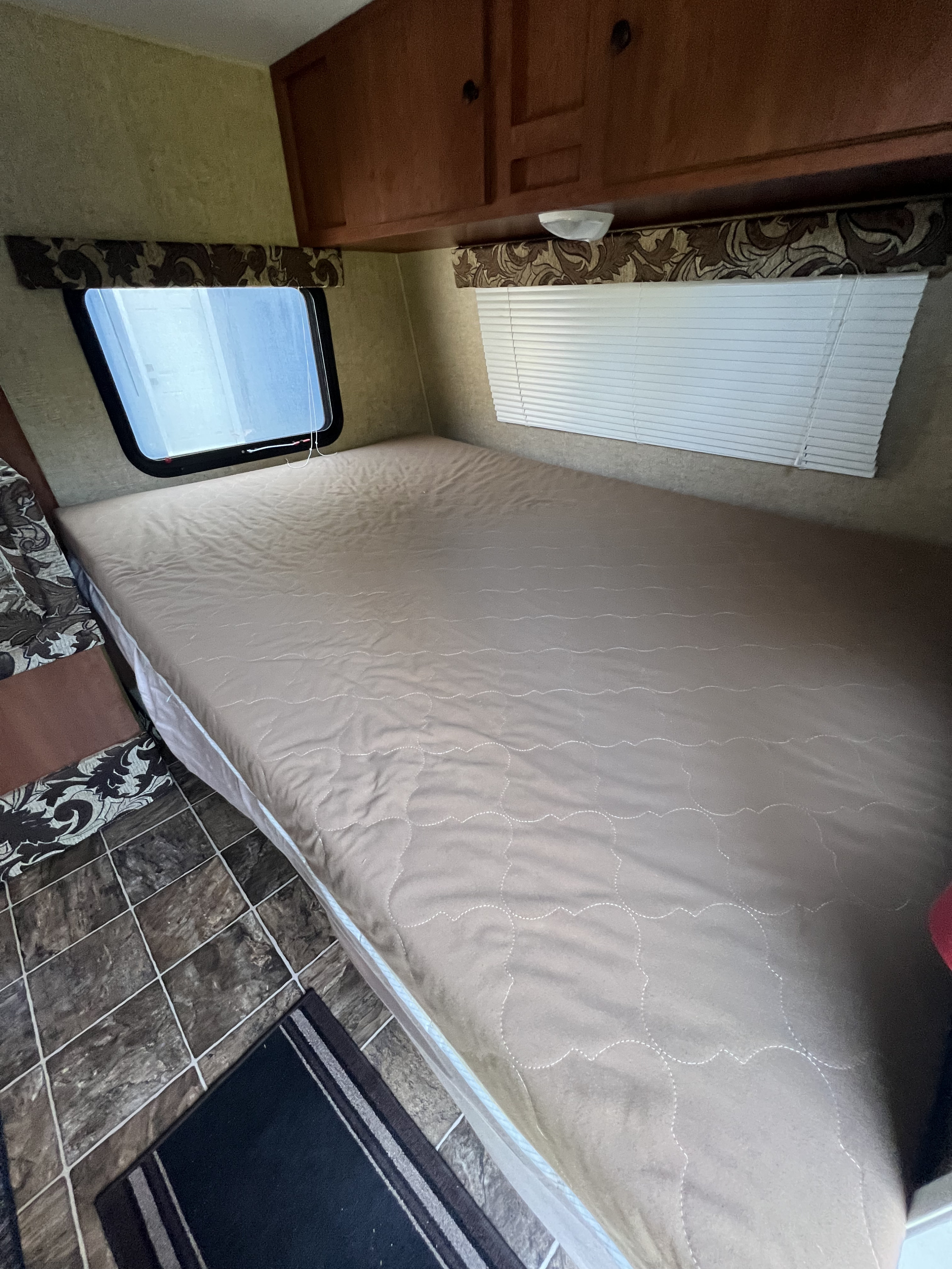 RV rental interior: bedroom with a tan quilted bed, two windows, wood cabinets, and tiled floor with a rug.. Sportsmen Classic 2014