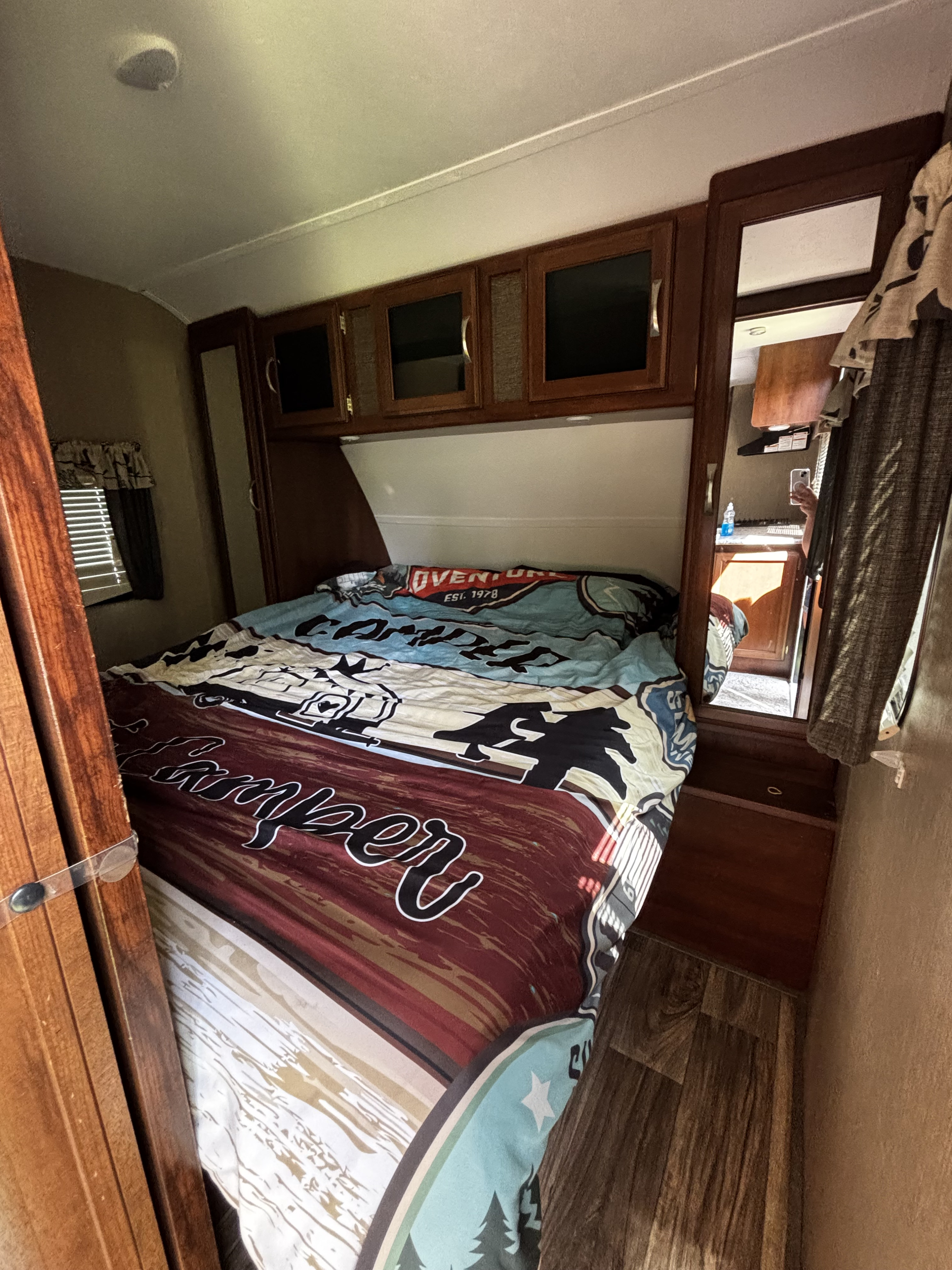 Cozy RV rental bedroom, featuring a large bed with a 'Camper Adventure' comforter, storage, and a kitchen reflection.. Keystone RV Passport Grand Touring 2016