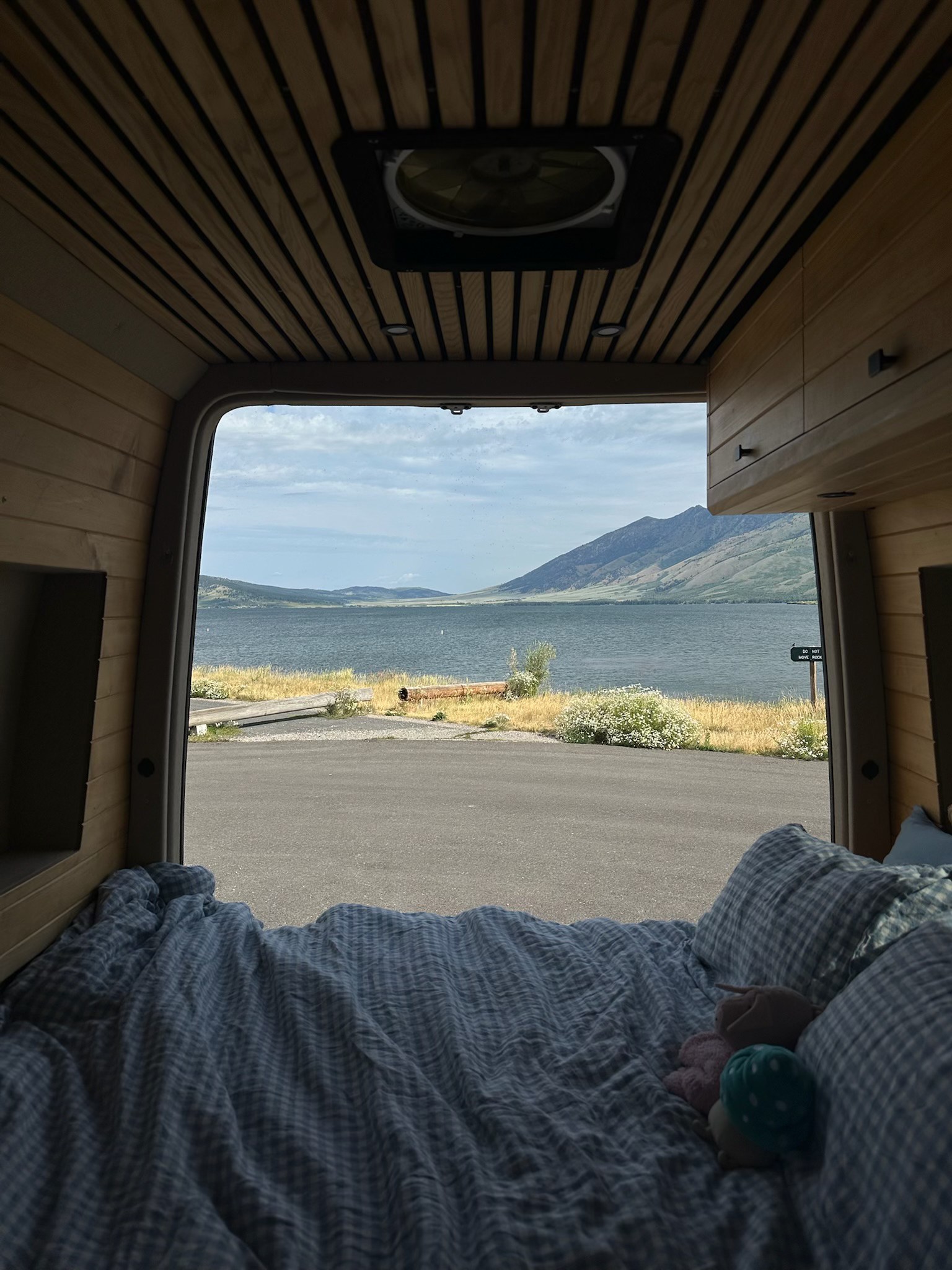 Enjoy the serene view from an RV rental's rear bed, featuring a cozy wood-paneled interior, a lake, mountains, & asphalt road.. Vanspeed Mercedes Sprinter 2021