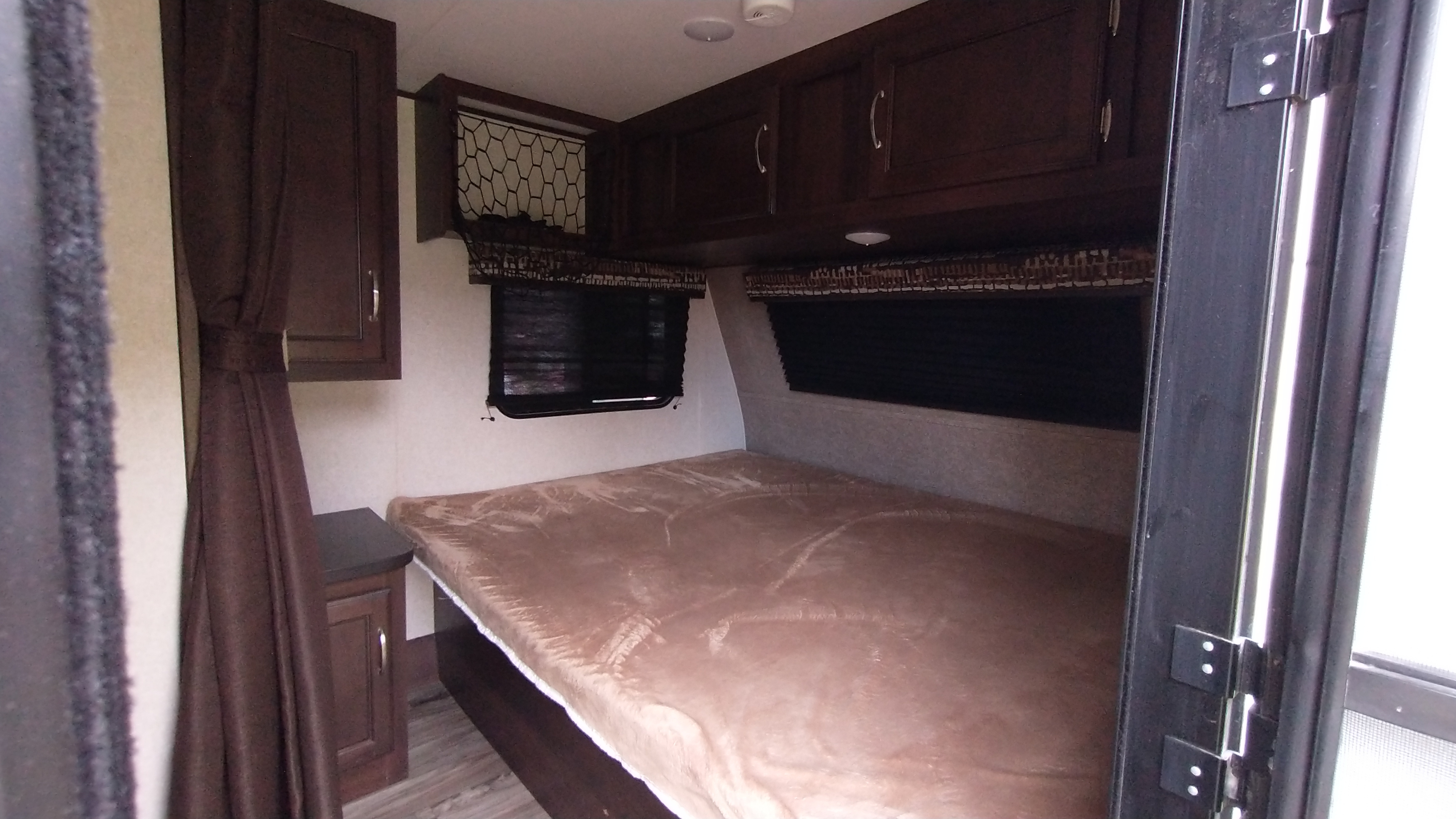 Interior of an rv rental shows a compact bedroom with a bed covered in a brown blanket, dark cabinets & windows.. Jayco Jay Flight SLX 2018