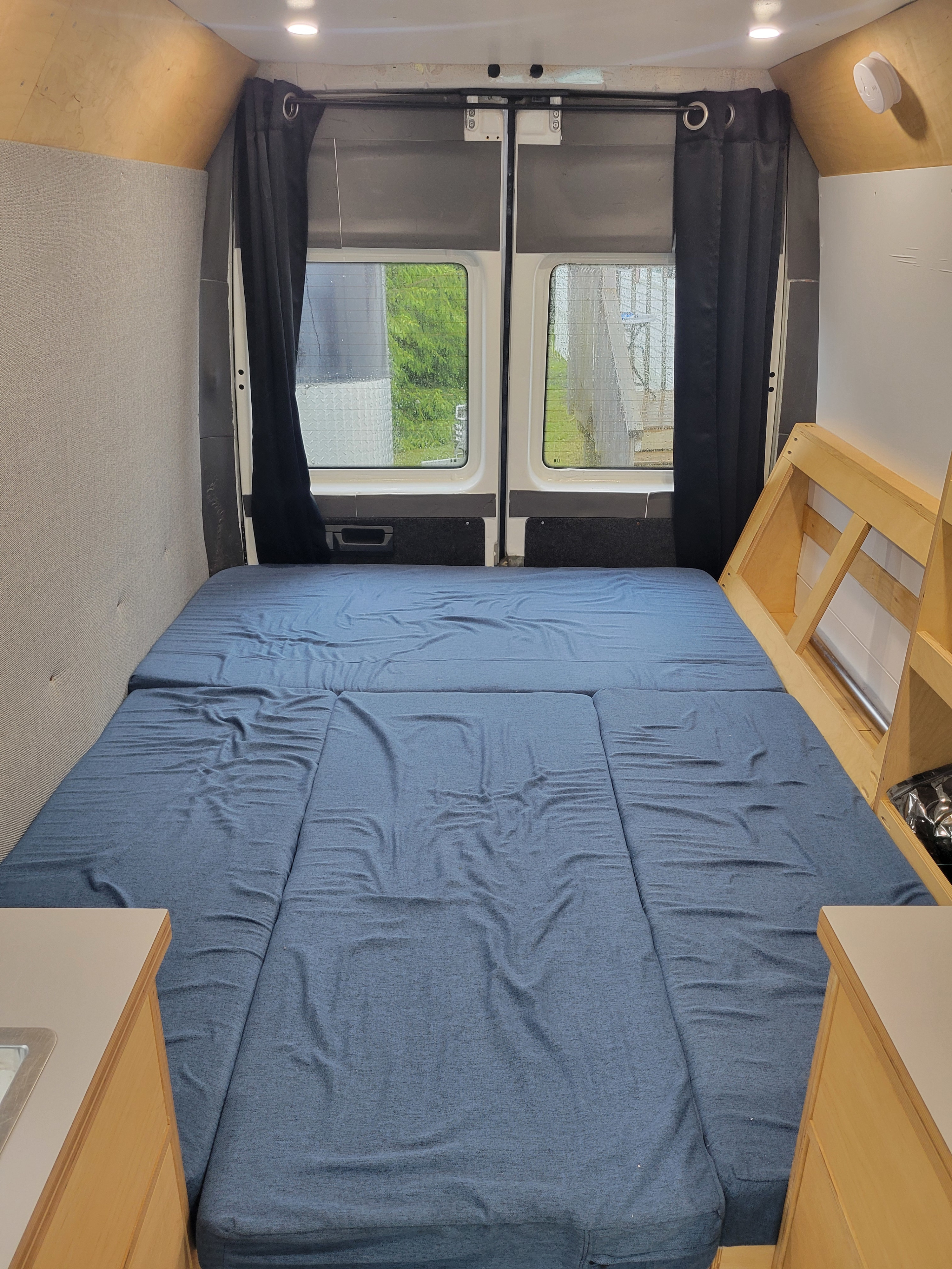 Inside an RV rental: full bed with blue mattress, rear windows, privacy curtains, and custom wood details.. Mercedes Sprinter 2006