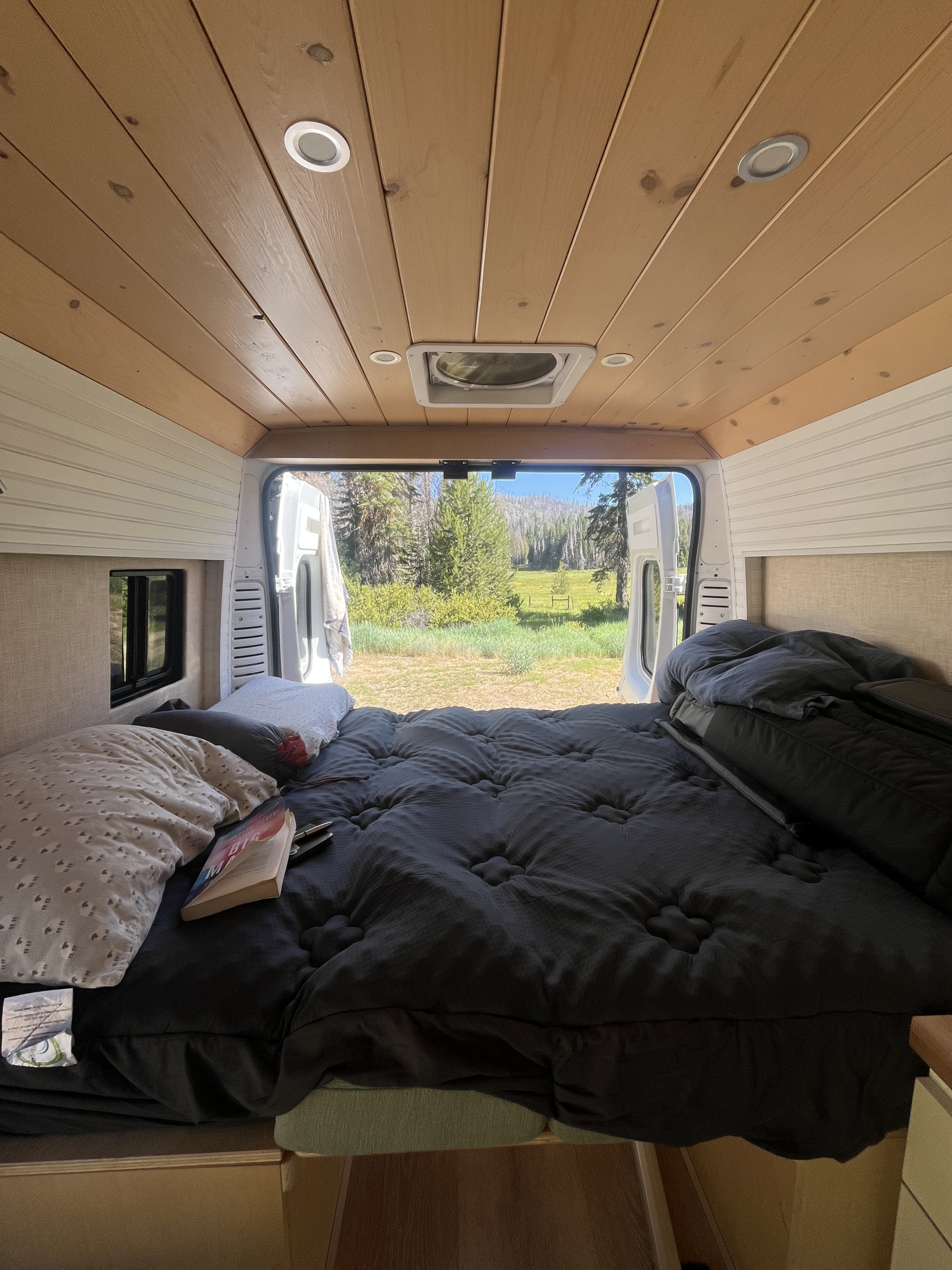 Cozy RV rental bed overlooks a serene forest and field through open rear doors. Perfect for a peaceful retreat.. Dodge Ram 1500 2021