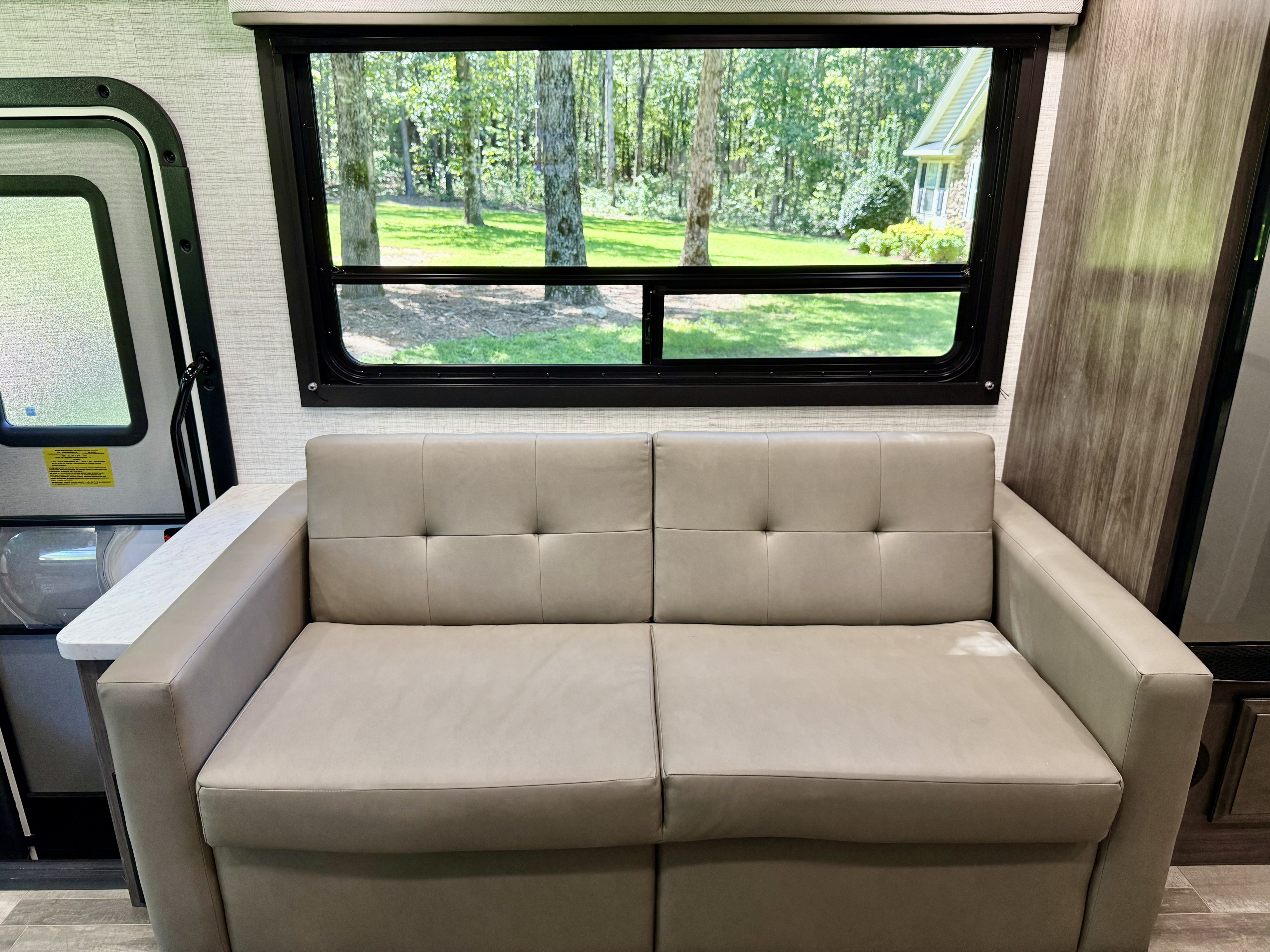 Cozy RV rental interior with a comfy tan sofa, large window revealing a lush outdoor view, and side door access.. Entegra Coach Vison 27ASE 2025