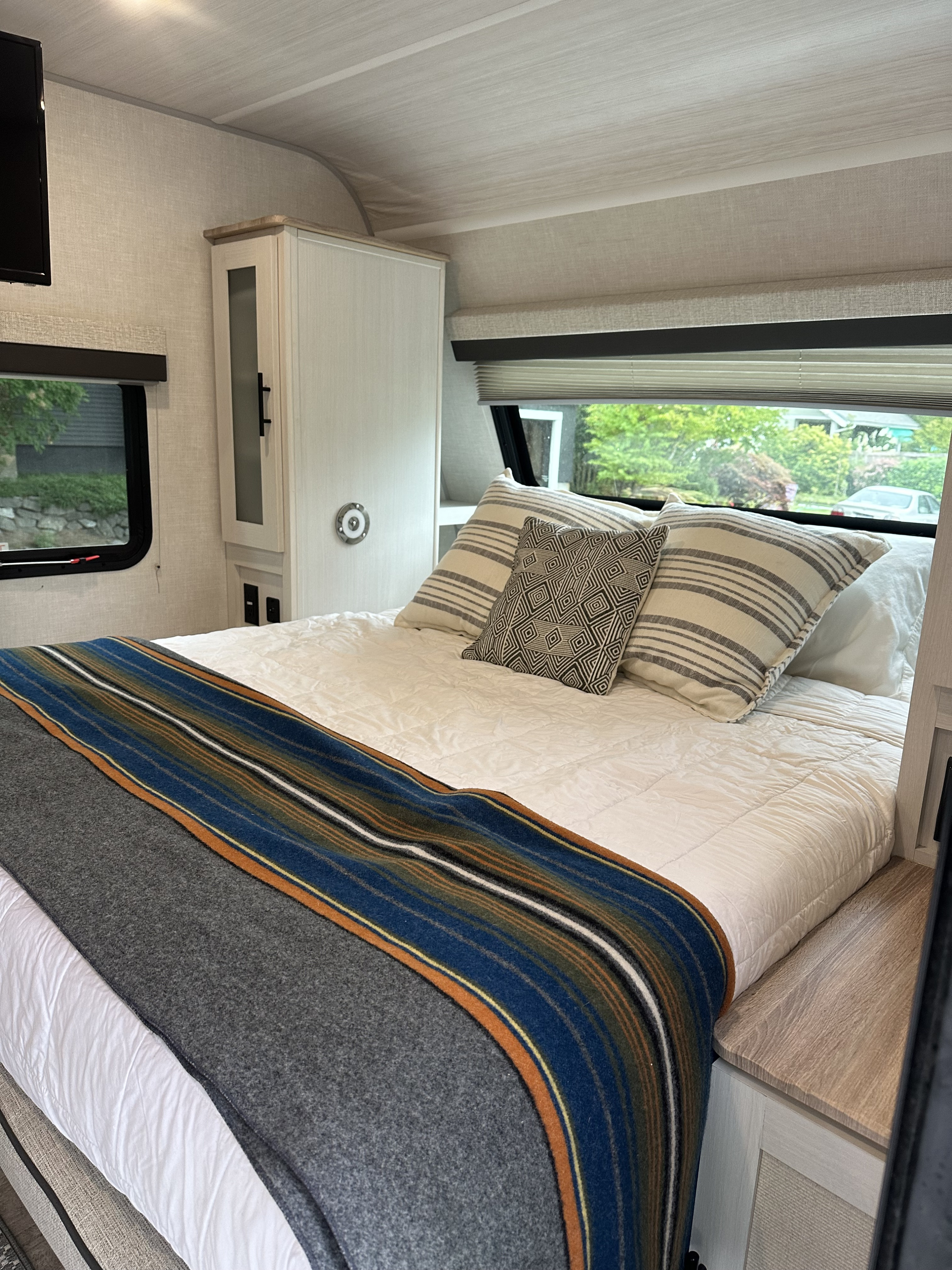 Comfortable RV rental bedroom: cozy bed, striped blanket, storage, and windows showing outdoor greenery.. No Boundaries 19 2023