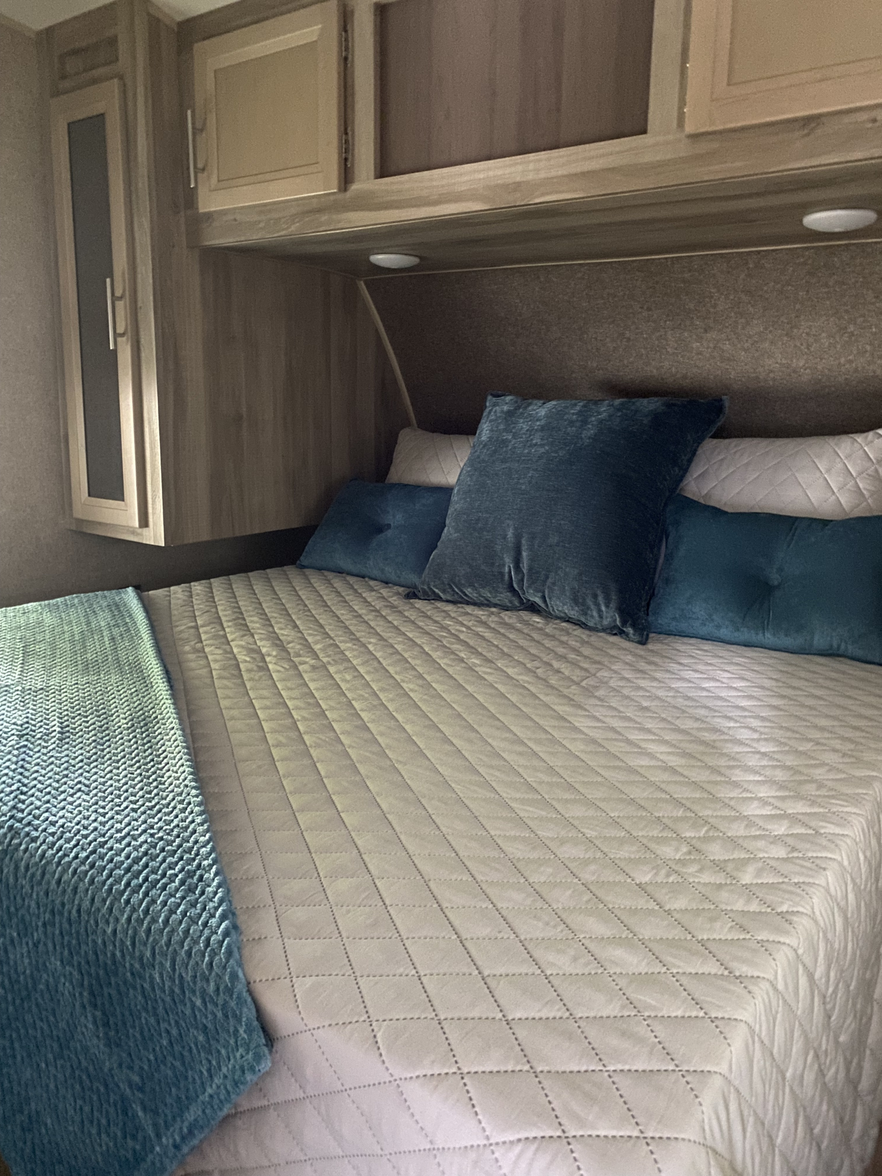 Inside an RV rental: a neatly made bed with grey quilted bedding, blue pillows, and built-in wood storage cabinets.. Forest River Coachmen Catalina Legacy 2019