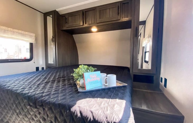 Cozy RV rental bedroom with a dark quilted bed, overhead cabinets, a bedside mirror, and a tray with coffee mugs.. Forest River Shasta Oasis 2021