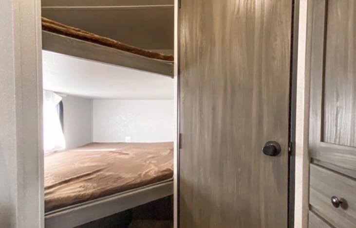 Inside an RV rental, showing compact bunk beds with brown bedding on the left. A wooden door with a dark knob is on the right.. Forest River Shasta Oasis 2021