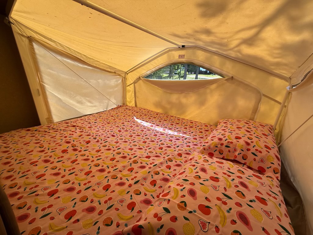 Cozy RV rental bed inside a pop-up camper, featuring fun fruit-patterned sheets and a window view of trees.. Forest River Rockwood Roo 2011