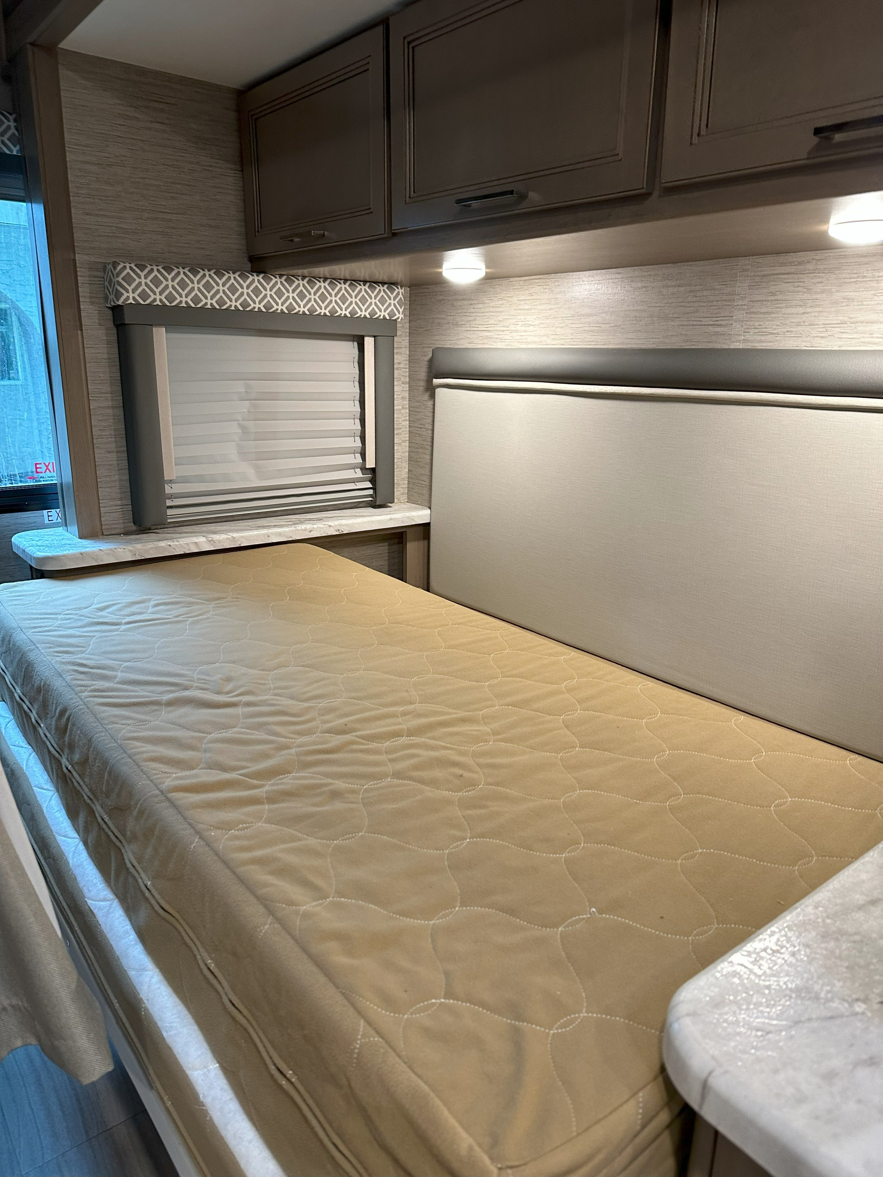 View inside an RV rental showing a quilted bed, window with blinds, overhead cabinets, and a padded backrest.. Thor Coleman 2022