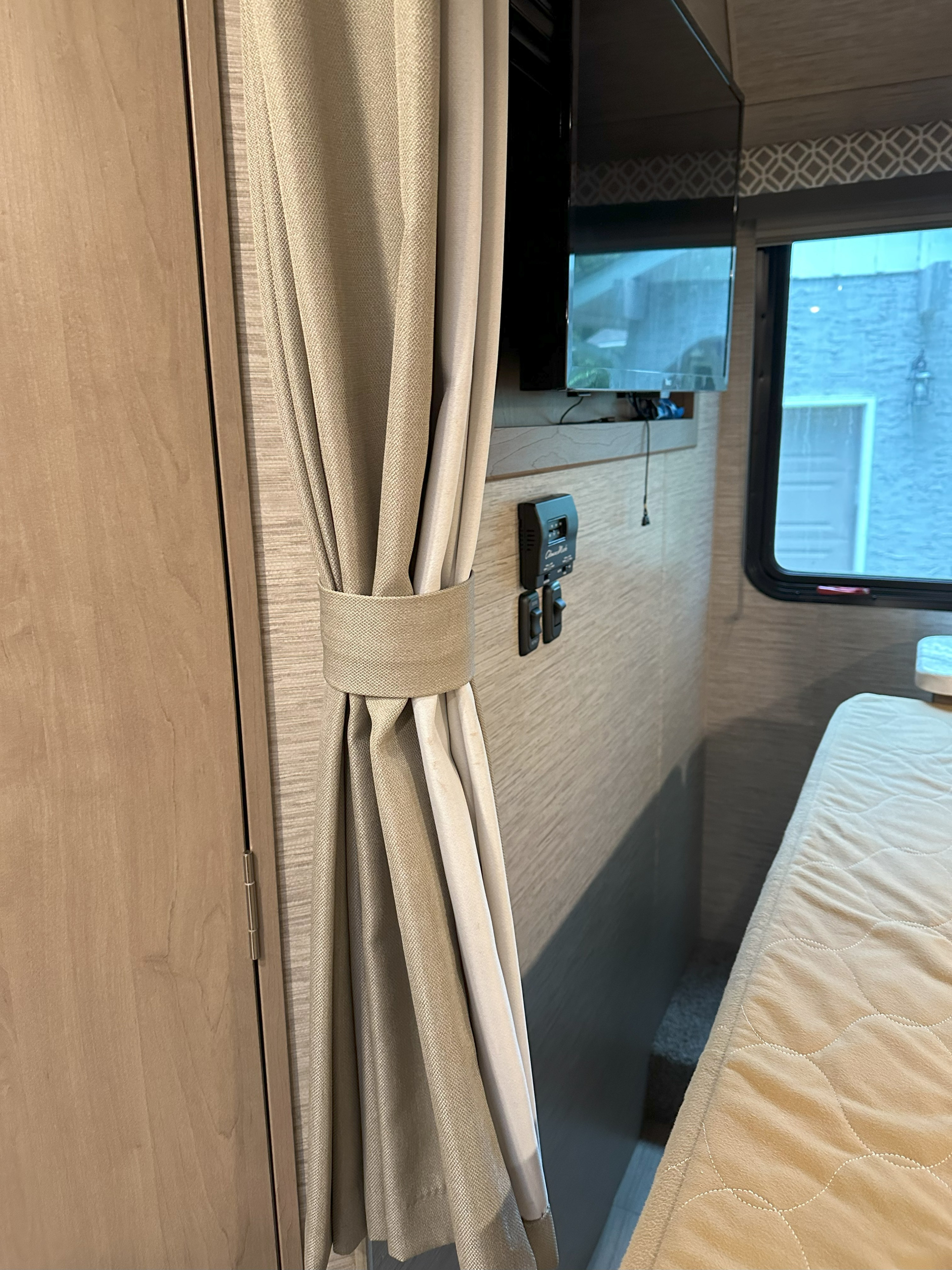 Cozy RV rental interior shows a tied-back curtain, wood cabinet, bed, wall-mounted TV & controls.. Thor Coleman 2022