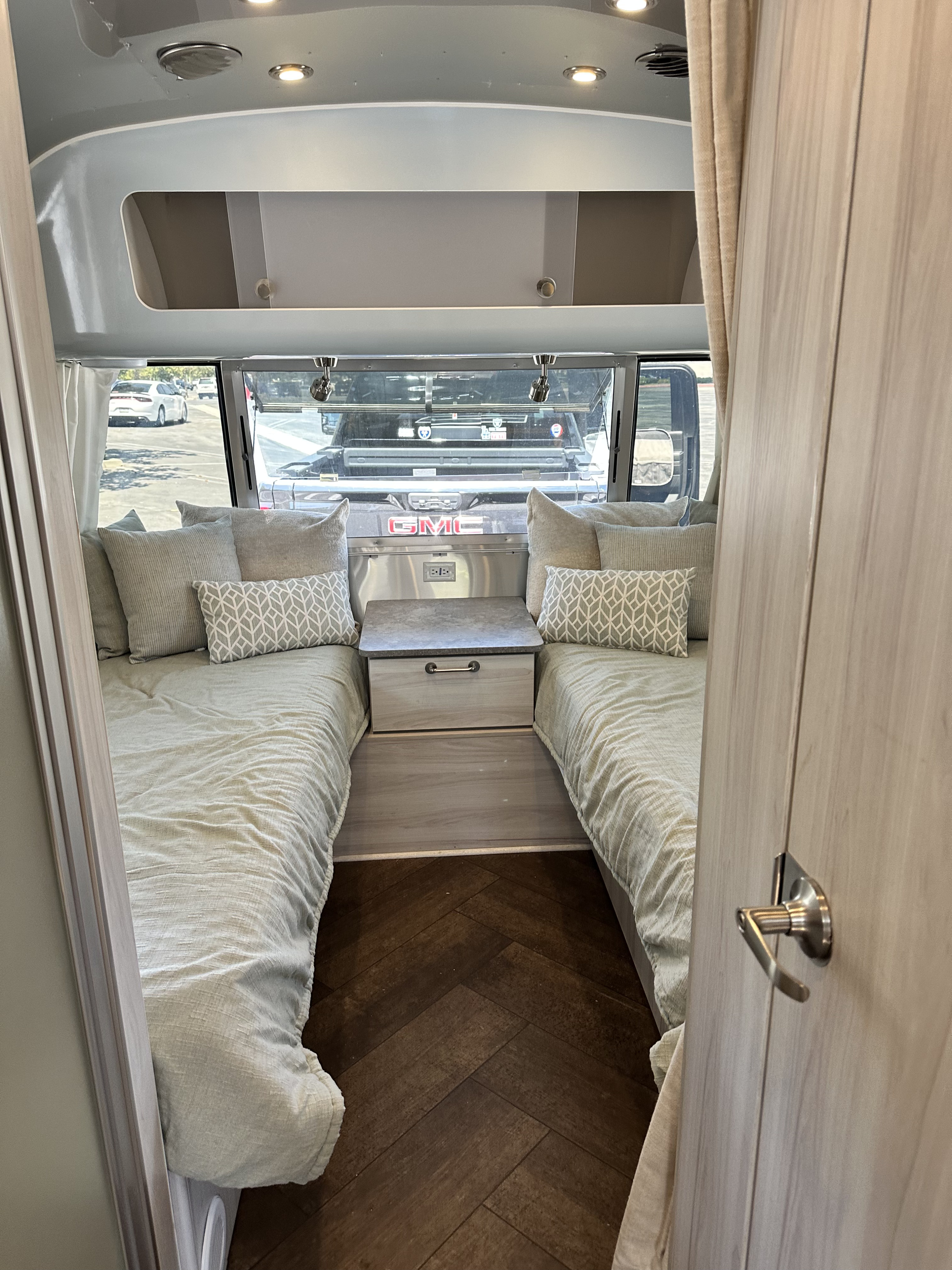 Cozy RV rental bedroom with two twin beds, a nightstand with power, overhead storage, and stylish herringbone flooring.. Airstream International 2023
