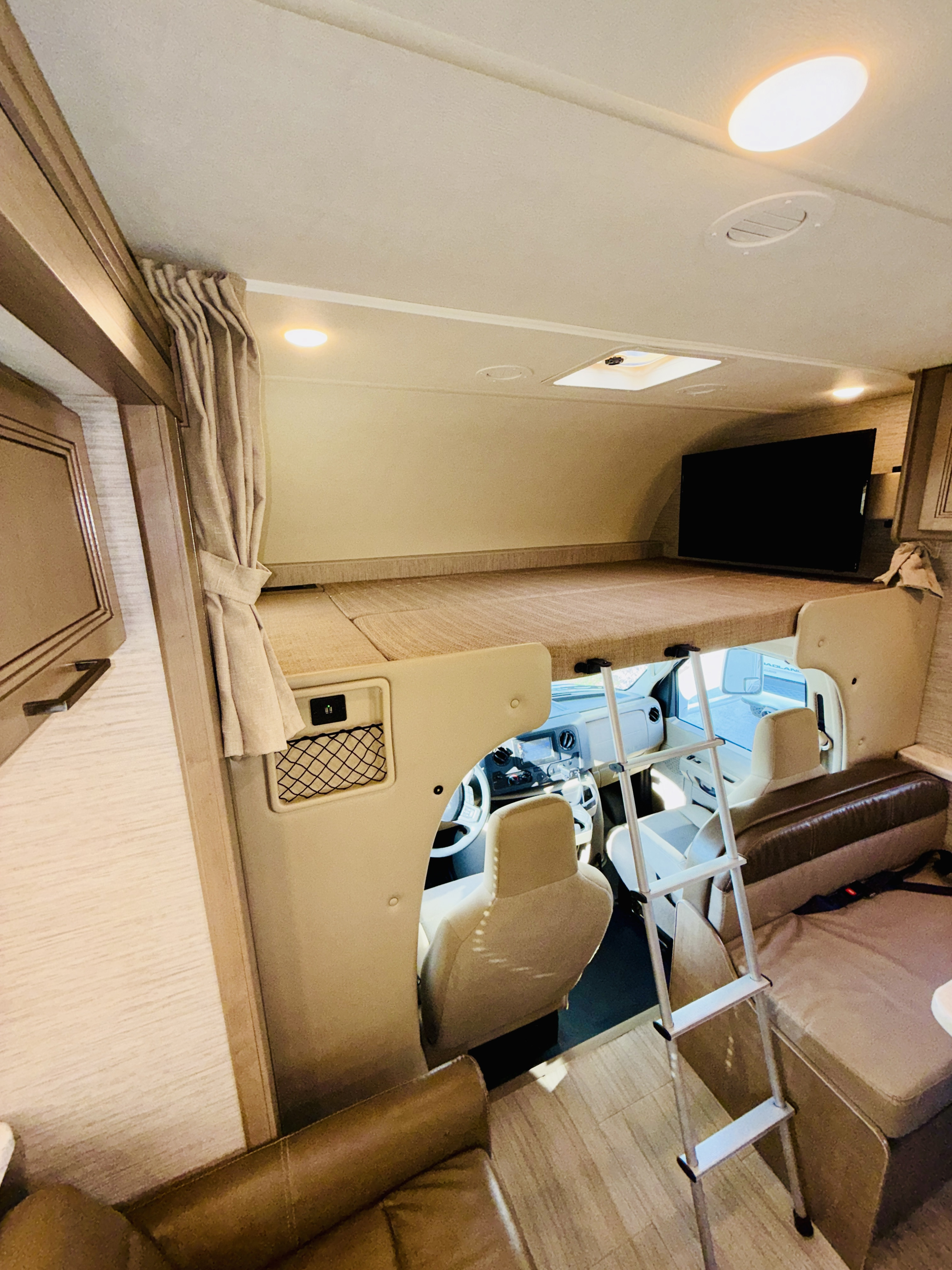 View inside an RV rental showing a cab-over bunk bed with a TV, a ladder for access, and the vehicle's front cabin.. Thor Coleman 2021