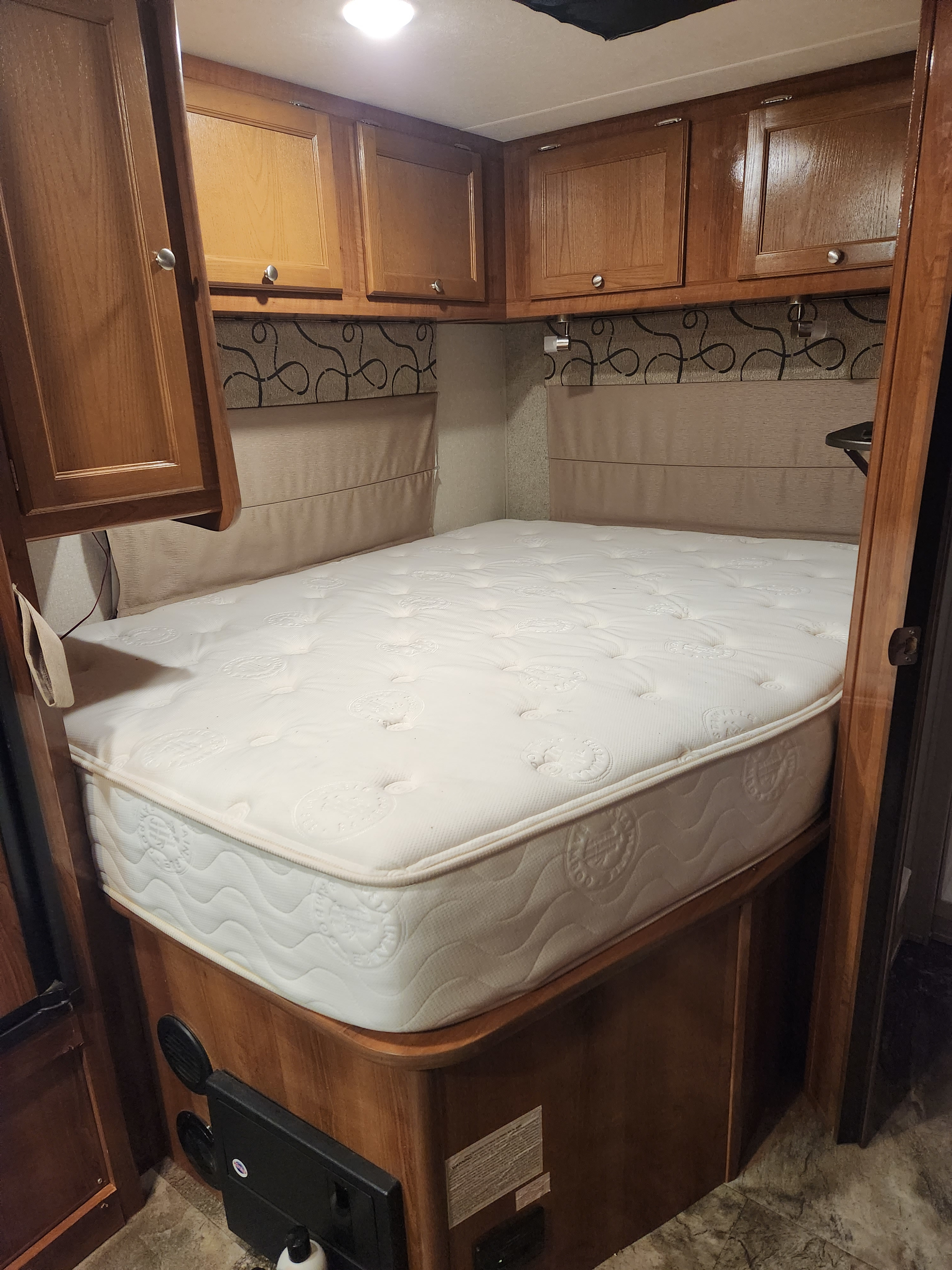 A clean, inviting RV rental bedroom with a white mattress, wood cabinetry, and patterned wall upholstery.. Coachmen Leprechaun 220QB 2019
