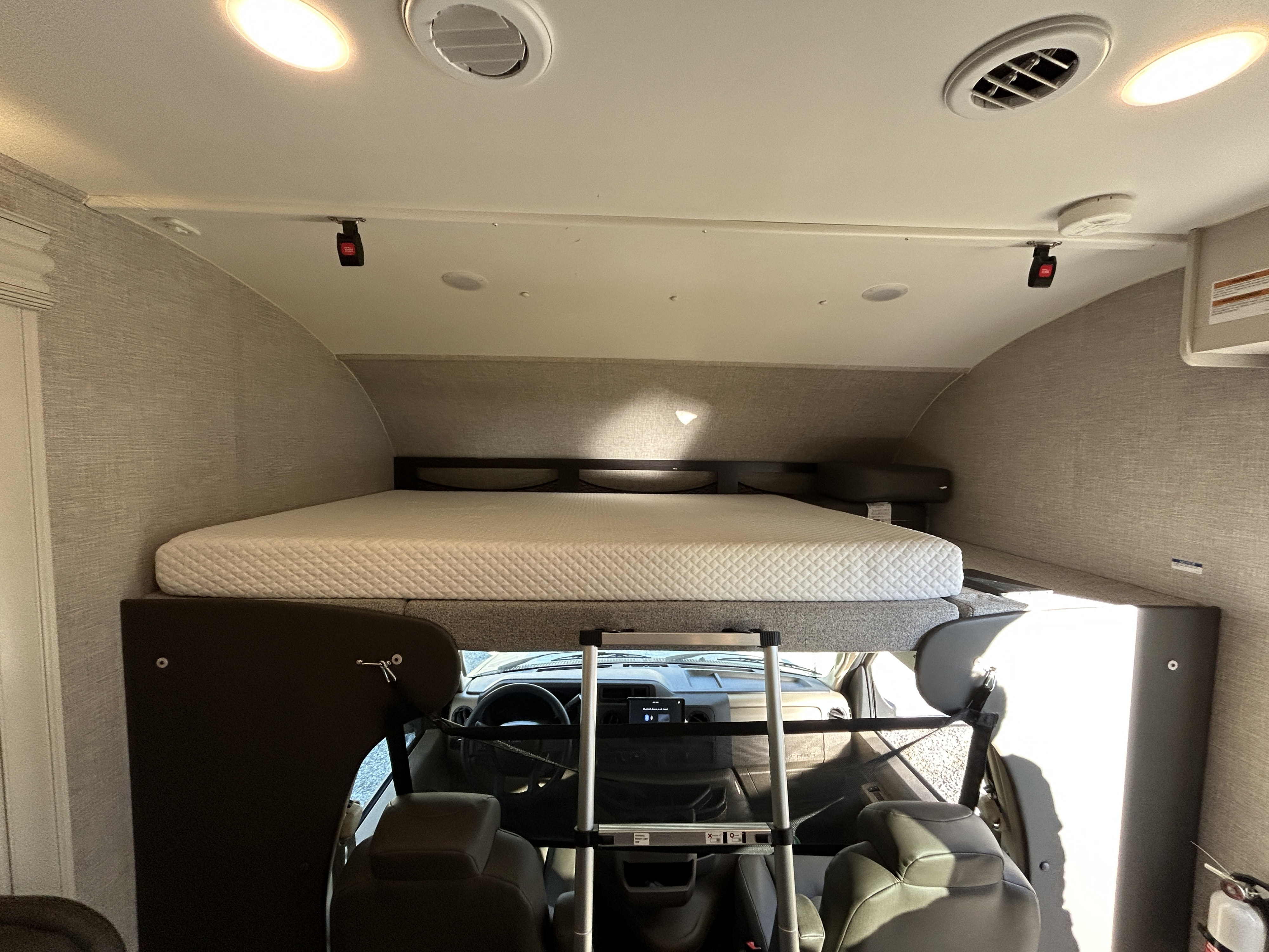 Inside an **rv rental**: a cozy cab-over bunk with a white mattress and ladder. The dashboard, overhead lights, and vents are visible below.. Jayco redhawk 2023