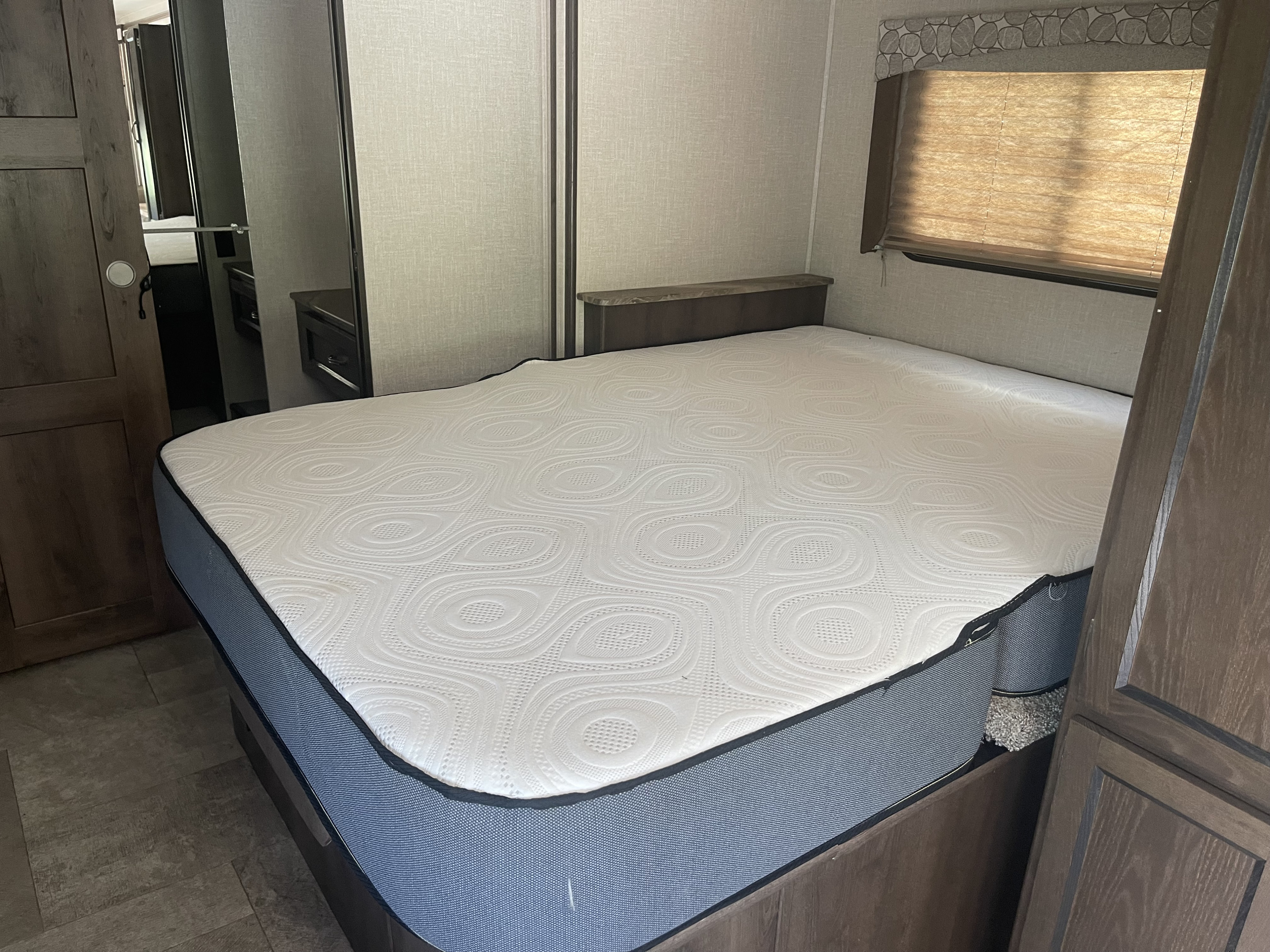 Experience comfort in this RV rental bedroom. A large patterned mattress, window with blinds, and wood accents create a cozy travel space.. Forest River Coachmen Freelander 2018
