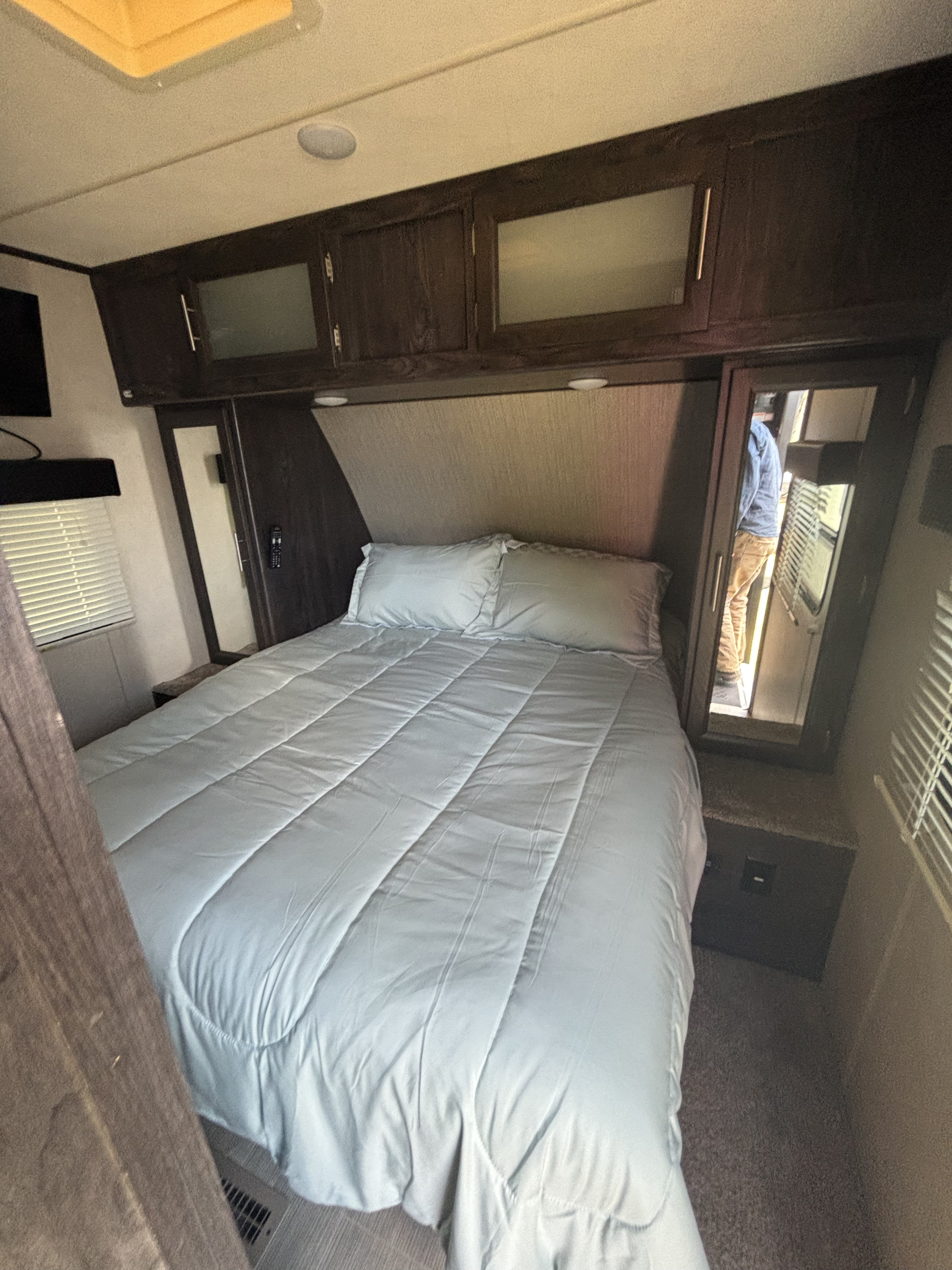 RV rental bedroom: A cozy camper interior with a bed, light blue bedding, overhead cabinets, window blinds, and carpeted floor.. Keystone RV 4YDT2332XJJ971604 2018