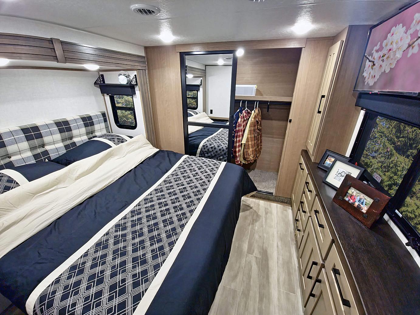 This RV rental bedroom offers a comfortable bed with plaid headboard, navy bedding, a closet, and dresser. Cozy travel accommodation.. K-Z Durango 2024