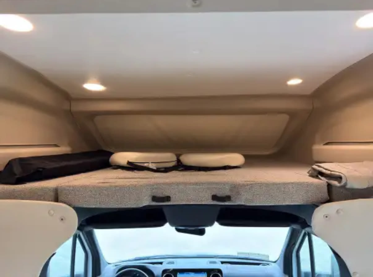 Inside an RV rental: view of the overhead bunk bed with cushions and blanket. The vehicle's dashboard is visible below, showing the front of this camper van.. Entegra Coach Qwest 2022