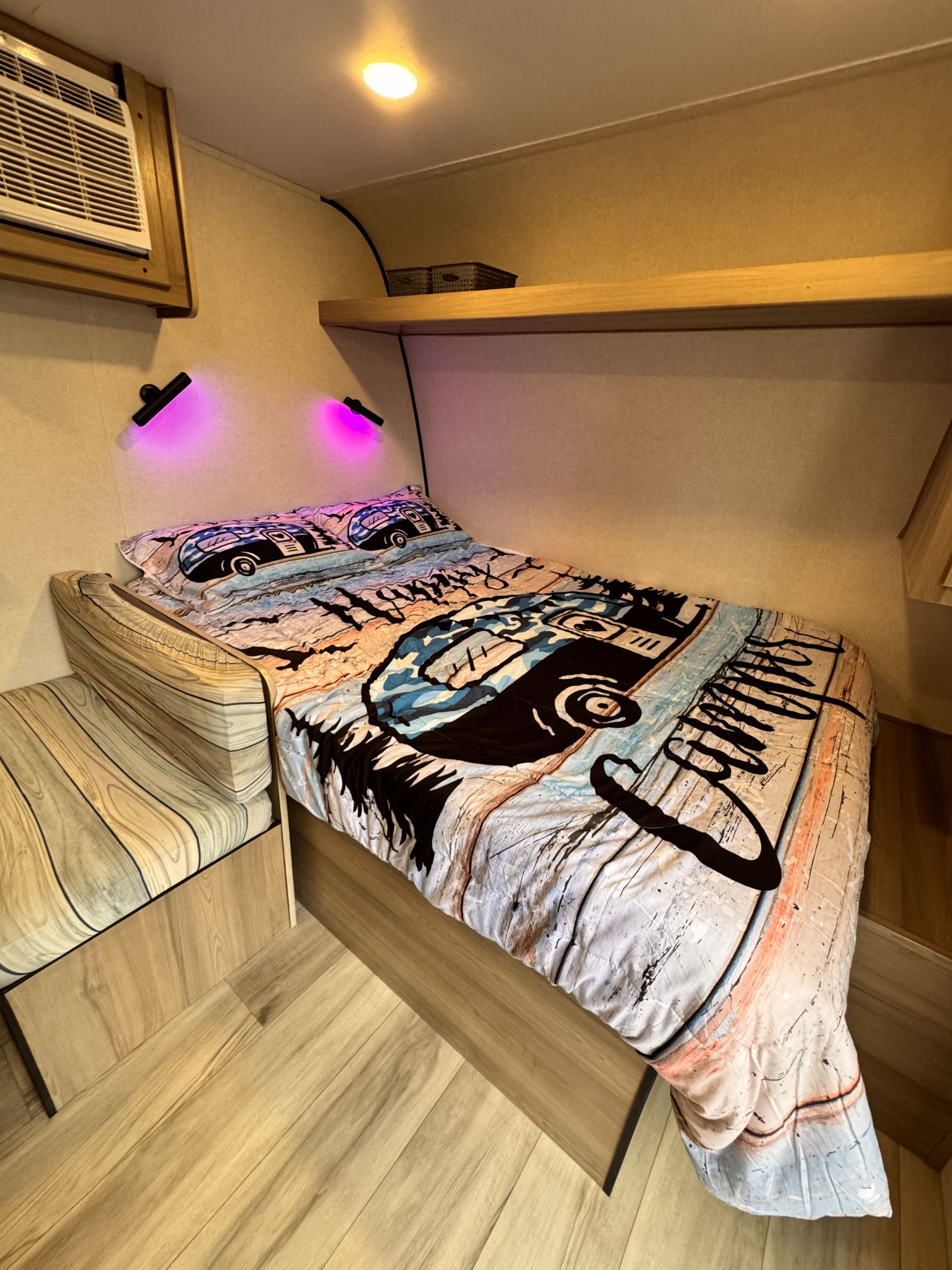 Cozy RV rental bedroom with colorful camper-themed bedding, purple lights, an AC unit, and a small sitting area.. Keystone Coleman 17B 2025